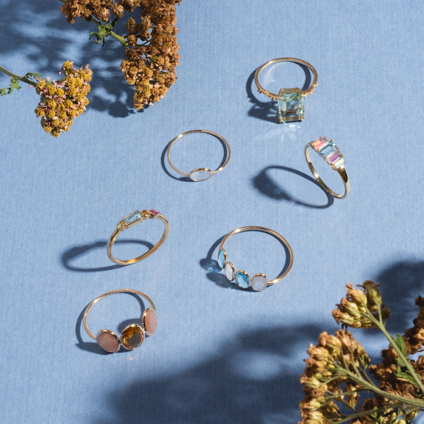 Seven delicate gold rings, featuring the Solid Gold Horizon Moonstone Ring, are displayed on a light blue surface and encircled by dried yellow flowers that cast soft shadows.