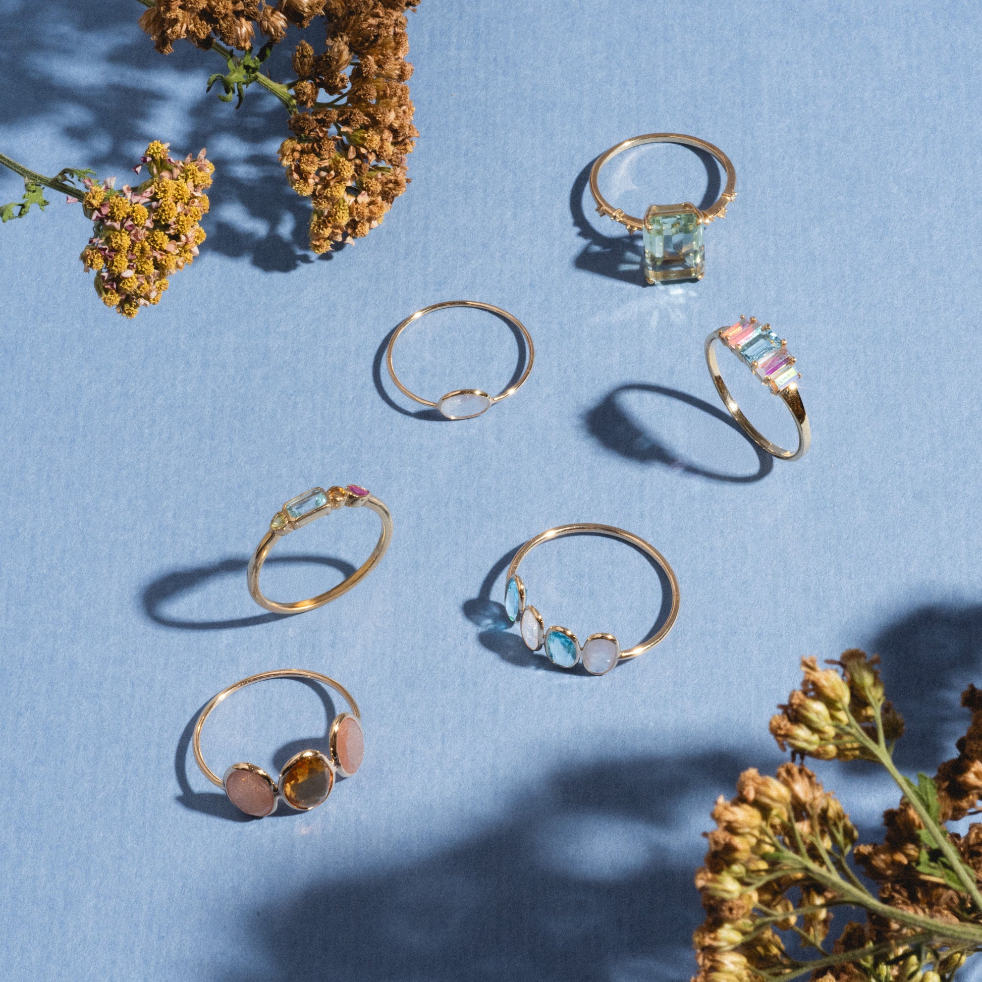 Seven delicate gold rings, featuring the Solid Gold Horizon Moonstone Ring, are displayed on a light blue surface and encircled by dried yellow flowers that cast soft shadows.