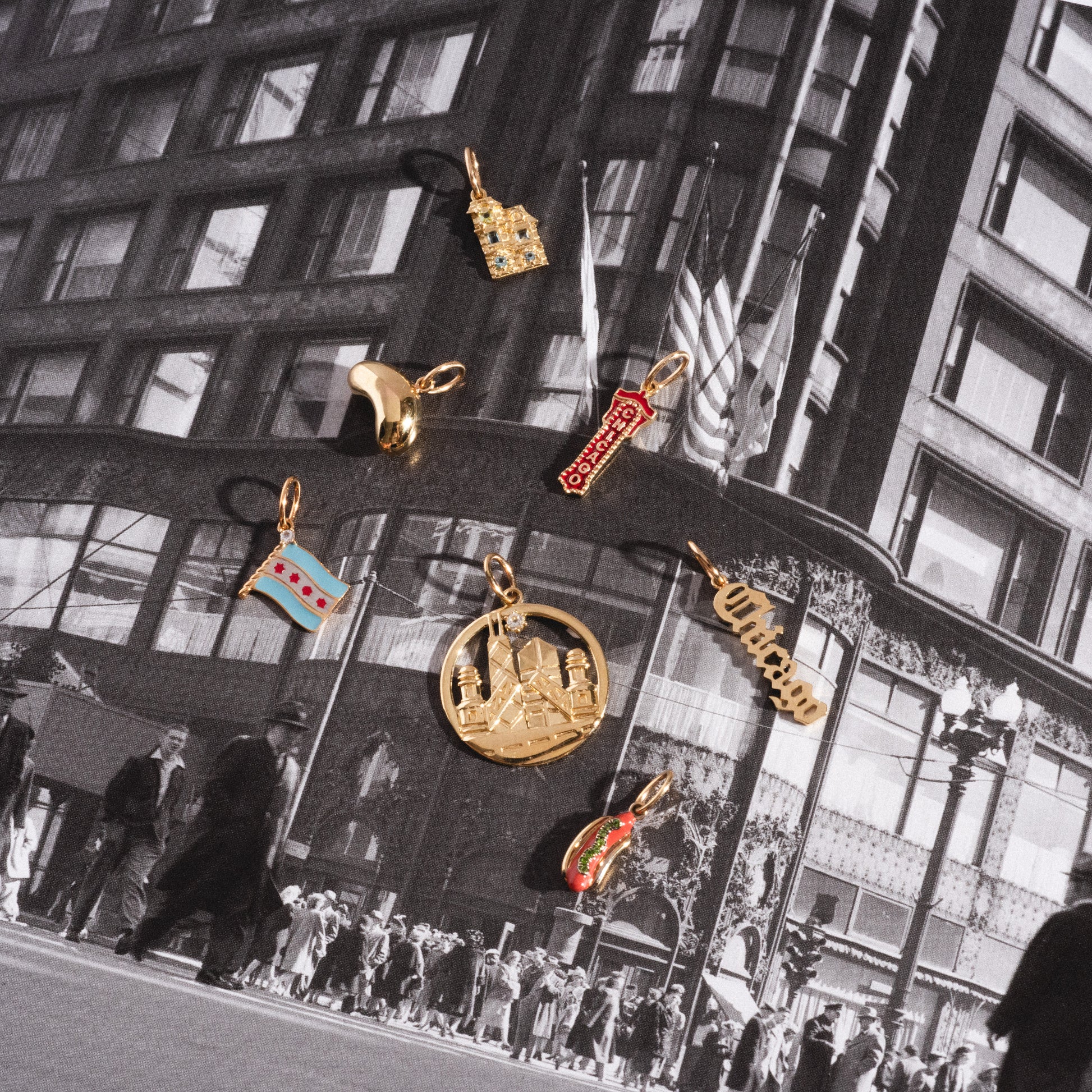 The Solid Gold Chicago Charm is displayed among seven colorful charms, arranged on a black-and-white vintage city street photo featuring a large building, U.S. flags, and pedestrians.