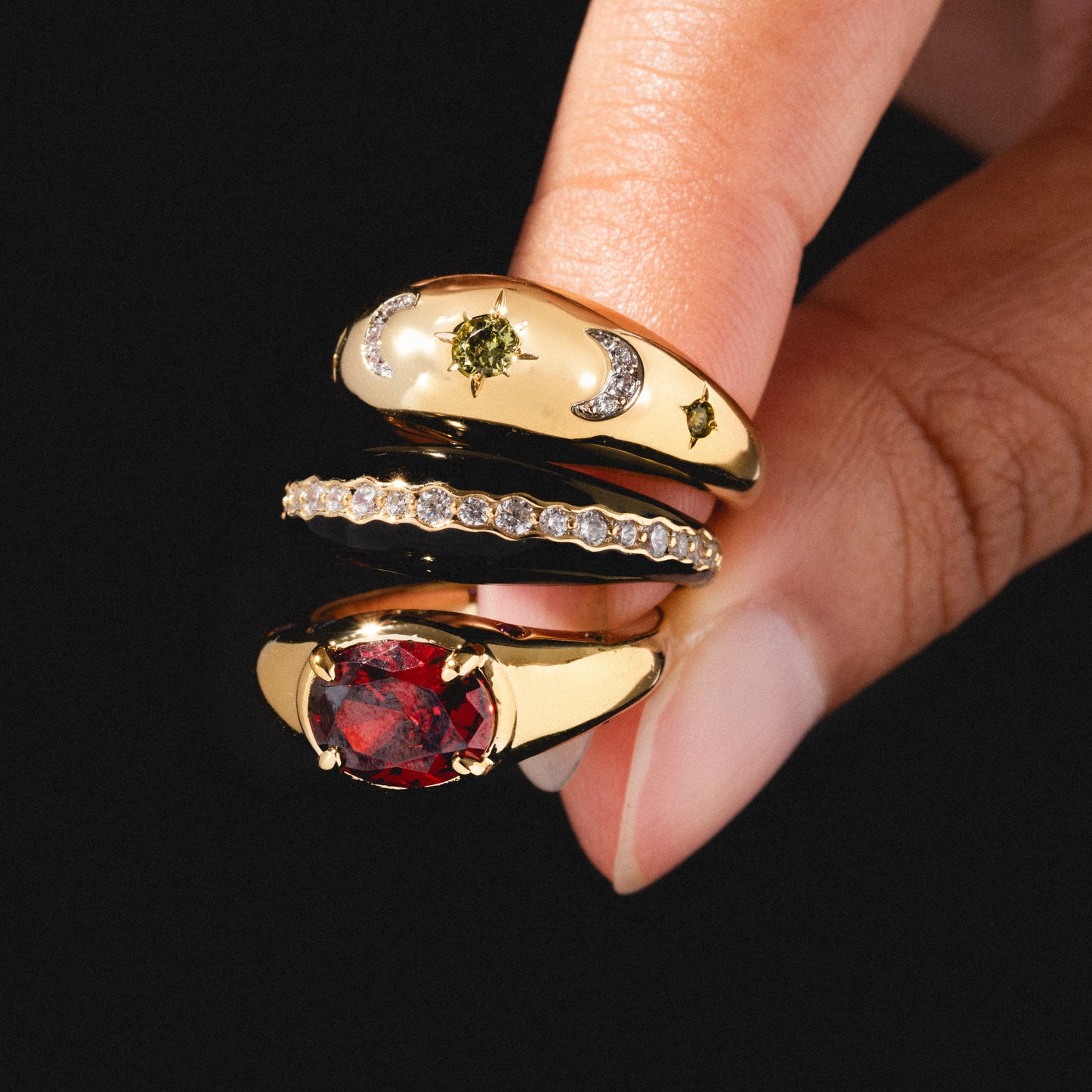 A hand displays the Free Spirit Black Enamel Ring with celestial designs and a green stone, stacked alongside a delicate CZ band with diamonds and a ring featuring a large oval red gemstone against a black background.