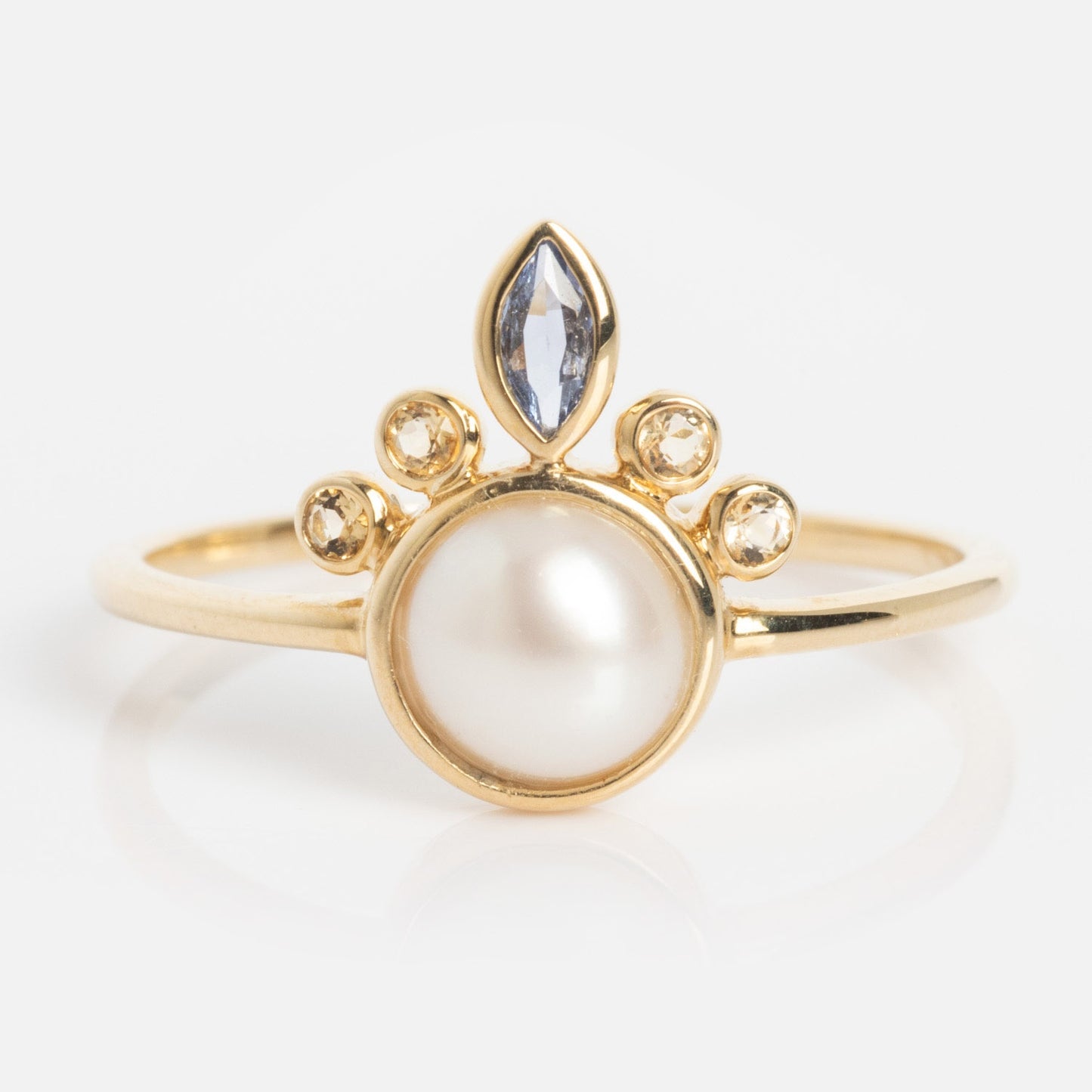 The Solid Gold Crowned Bezel Pearl Ring showcases a large central pearl, four small round diamonds above it, and a marquise-cut blue gemstone atop a thin gold band.