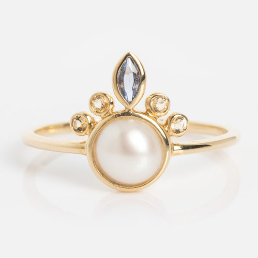 The Solid Gold Crowned Bezel Pearl Ring showcases a large central pearl, four small round diamonds above it, and a marquise-cut blue gemstone atop a thin gold band.