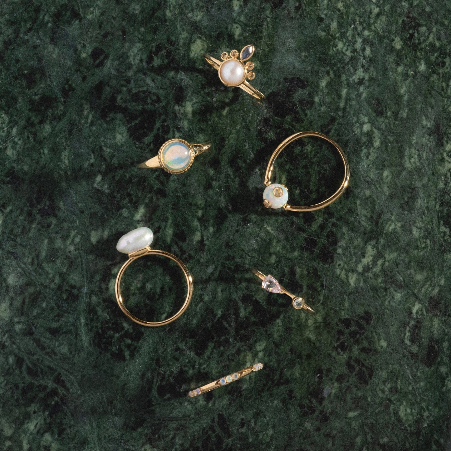 The Solid Gold Gem Drop Opal Spinner Ring features opals and sparkling stones, displayed on a dark green marble surface among other unique gold rings.