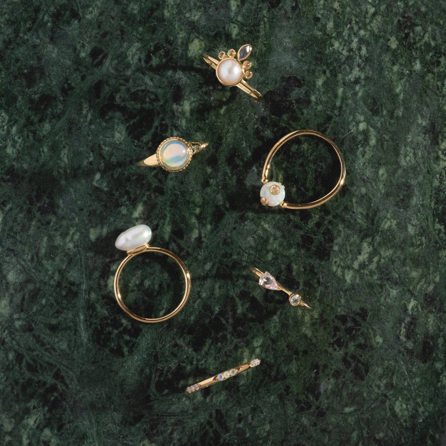 The Solid Gold Gem Drop Opal Spinner Ring features opals and sparkling stones, displayed on a dark green marble surface among other unique gold rings.