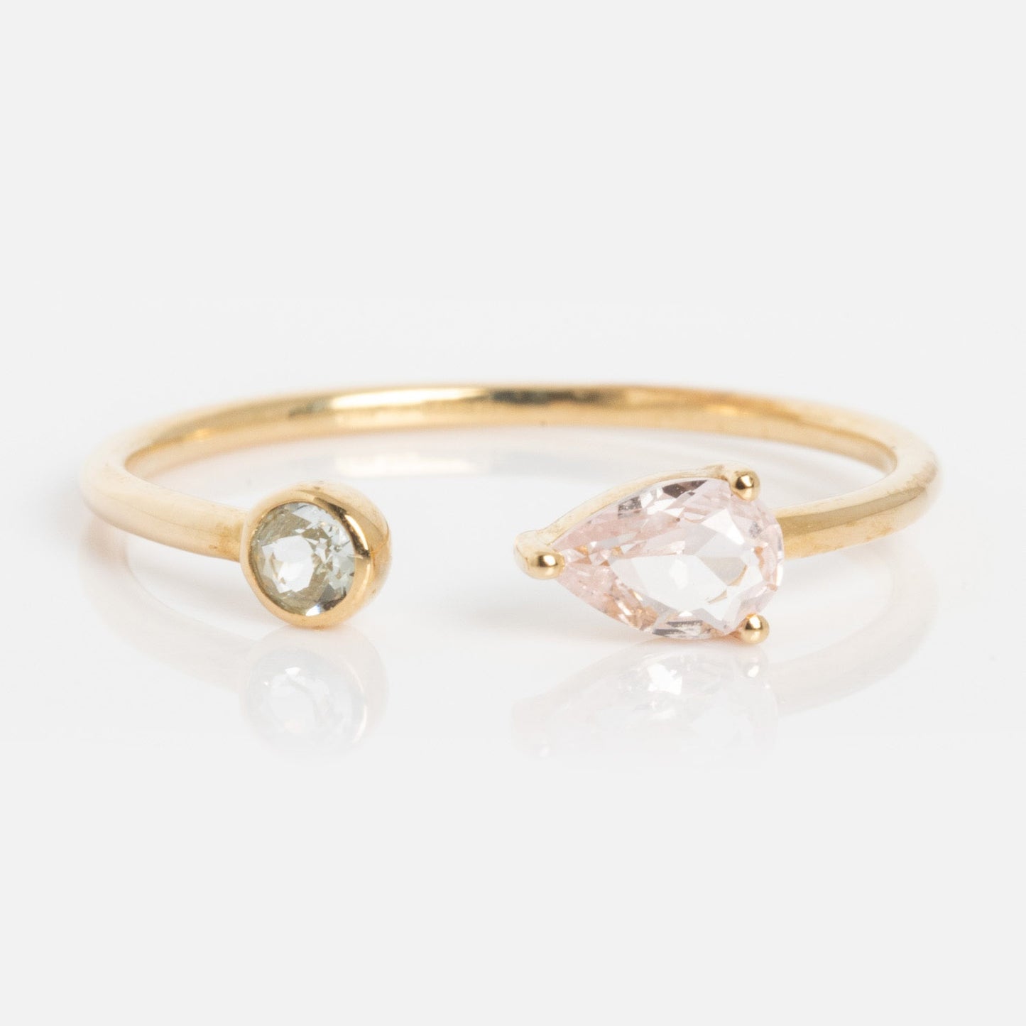 The Solid Gold Sapphire and Aquamarine Open Front Ring features a delicate open band with a round clear aquamarine on one end and a pear-shaped pale pink sapphire on the other, set against a white background.