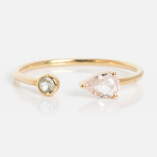The Solid Gold Sapphire and Aquamarine Open Front Ring features a delicate open band with a round clear aquamarine on one end and a pear-shaped pale pink sapphire on the other, set against a white background.
