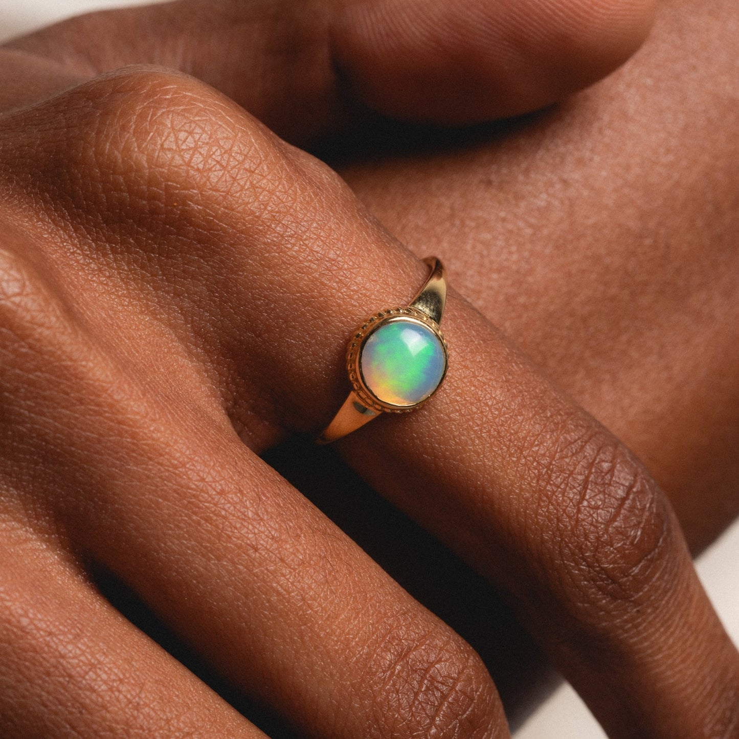 A close-up shows a hand wearing the Solid Gold Textured Opal Signet Ring, featuring an oval opal gemstone with iridescent colors. The ring sits on the finger, highlighting the opal's vibrant glow.