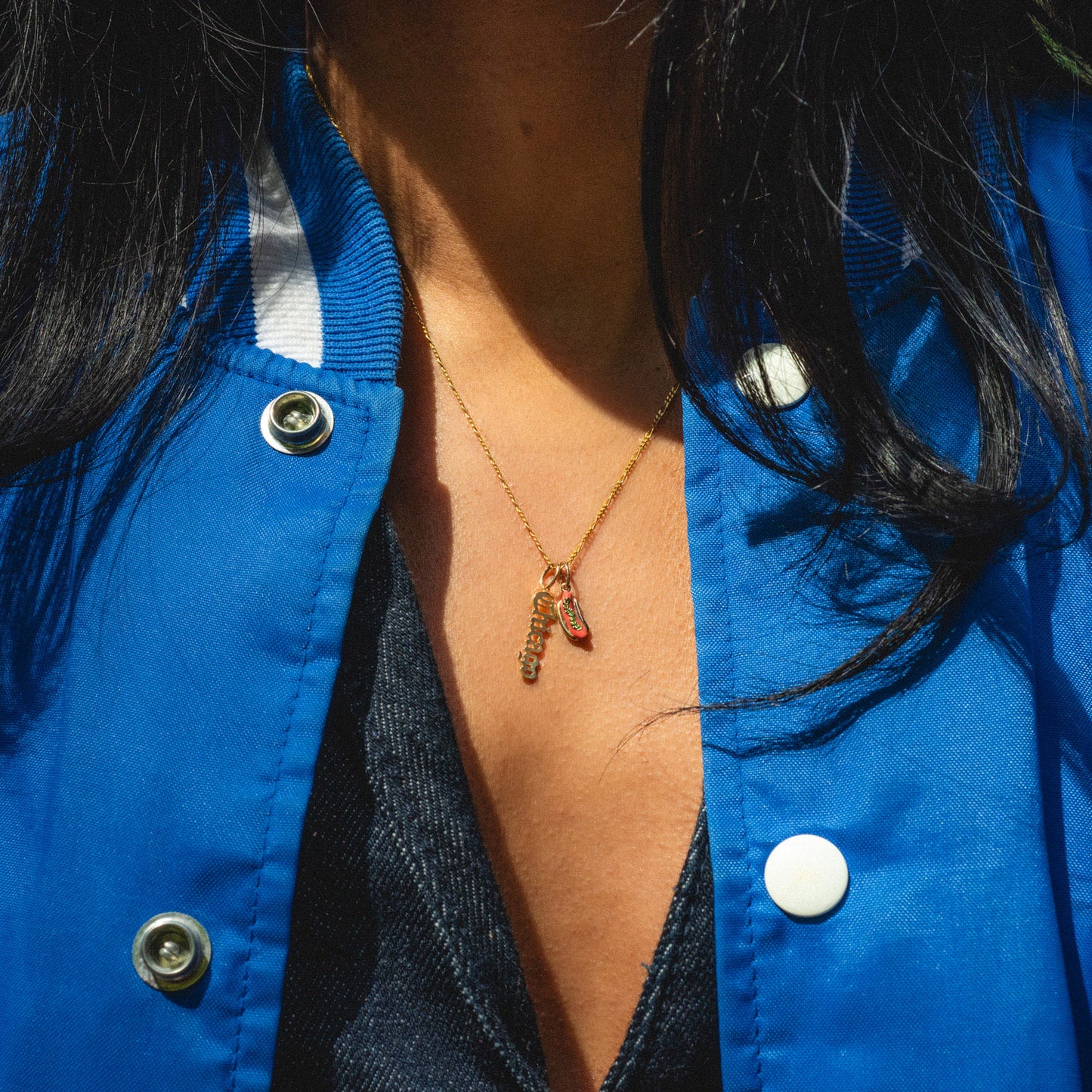 A person in a blue jacket over a dark top wears a gold necklace with two pendants—a sneaker-shaped charm and the Solid Gold Chicago Charm—around their neck.