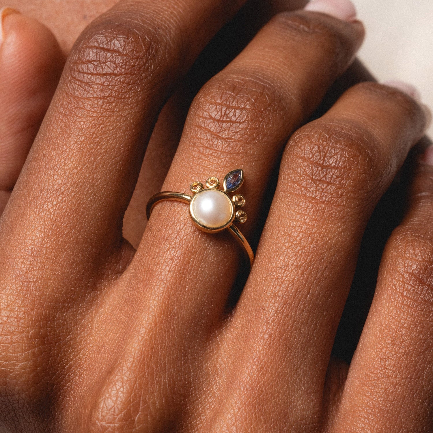 A close-up of a hand wearing the Solid Gold Crowned Bezel Pearl Ring, featuring a central pearl, three small round gems, and a marquise-shaped gem on top, resting against the fingers.