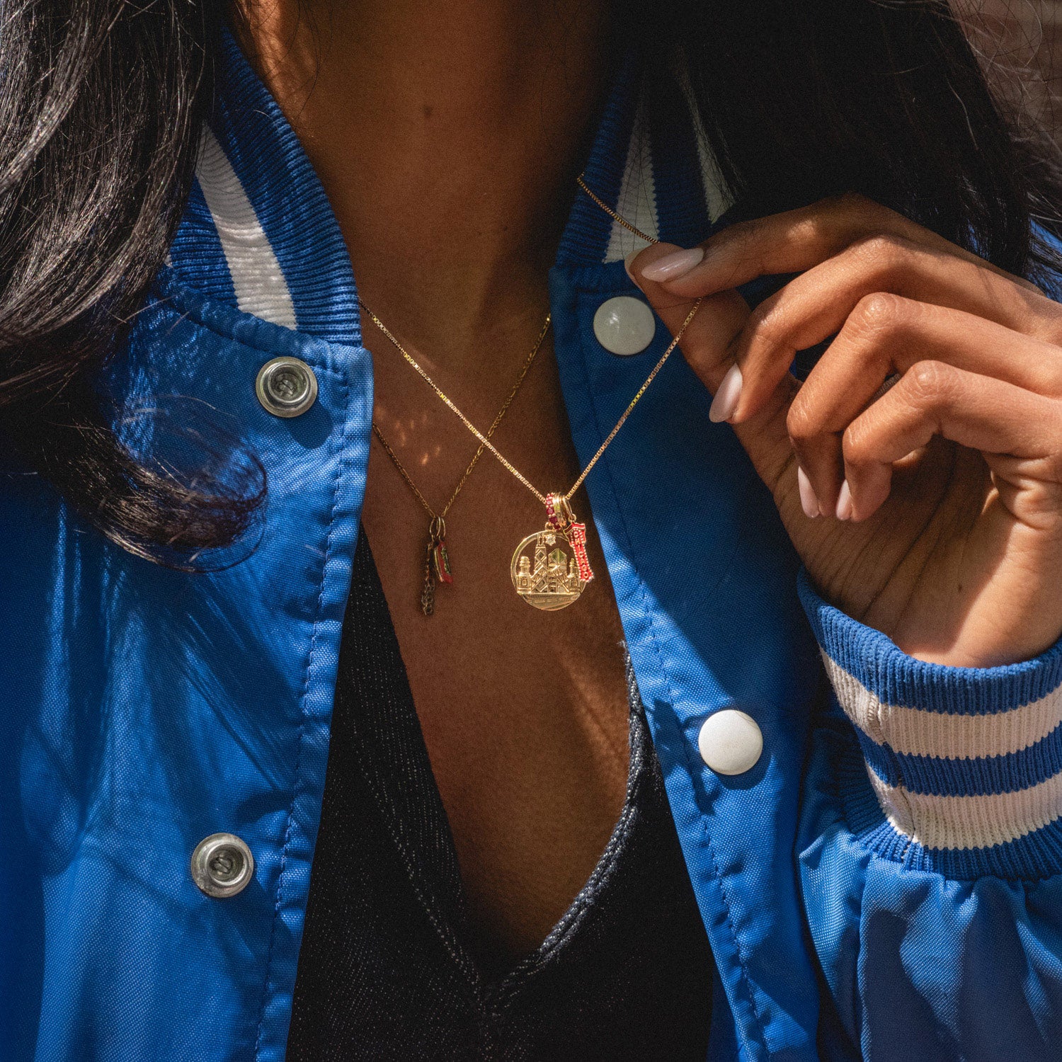 A woman in a blue varsity jacket showcases her jewelry, holding up a gold necklace with the Solid Gold Chicago Charm and another necklace featuring a round pendant.