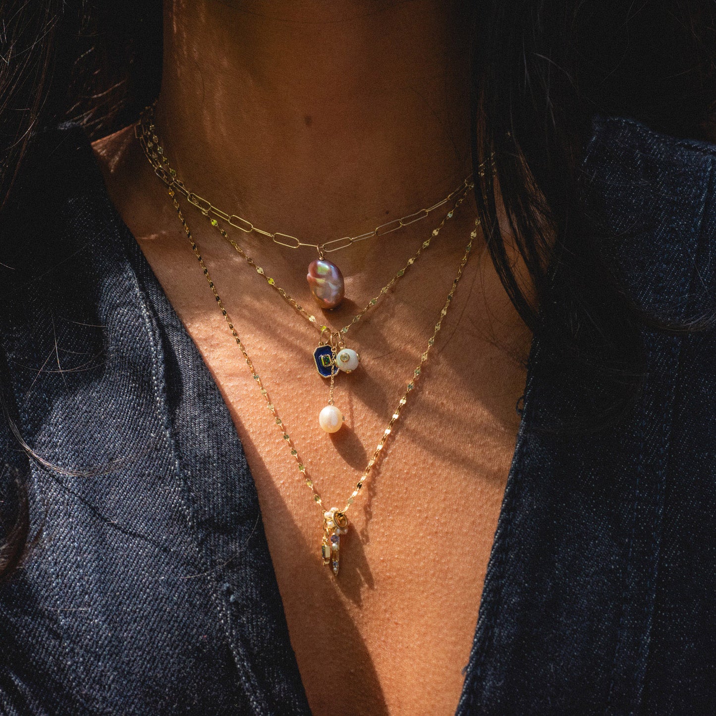 A person in a dark denim top wears three layered gold necklaces with unique charms—a pearl, a blue gem, and the Solid Gold Statement Charm—highlighted by sunlight. All are Local Eclectic exclusives.