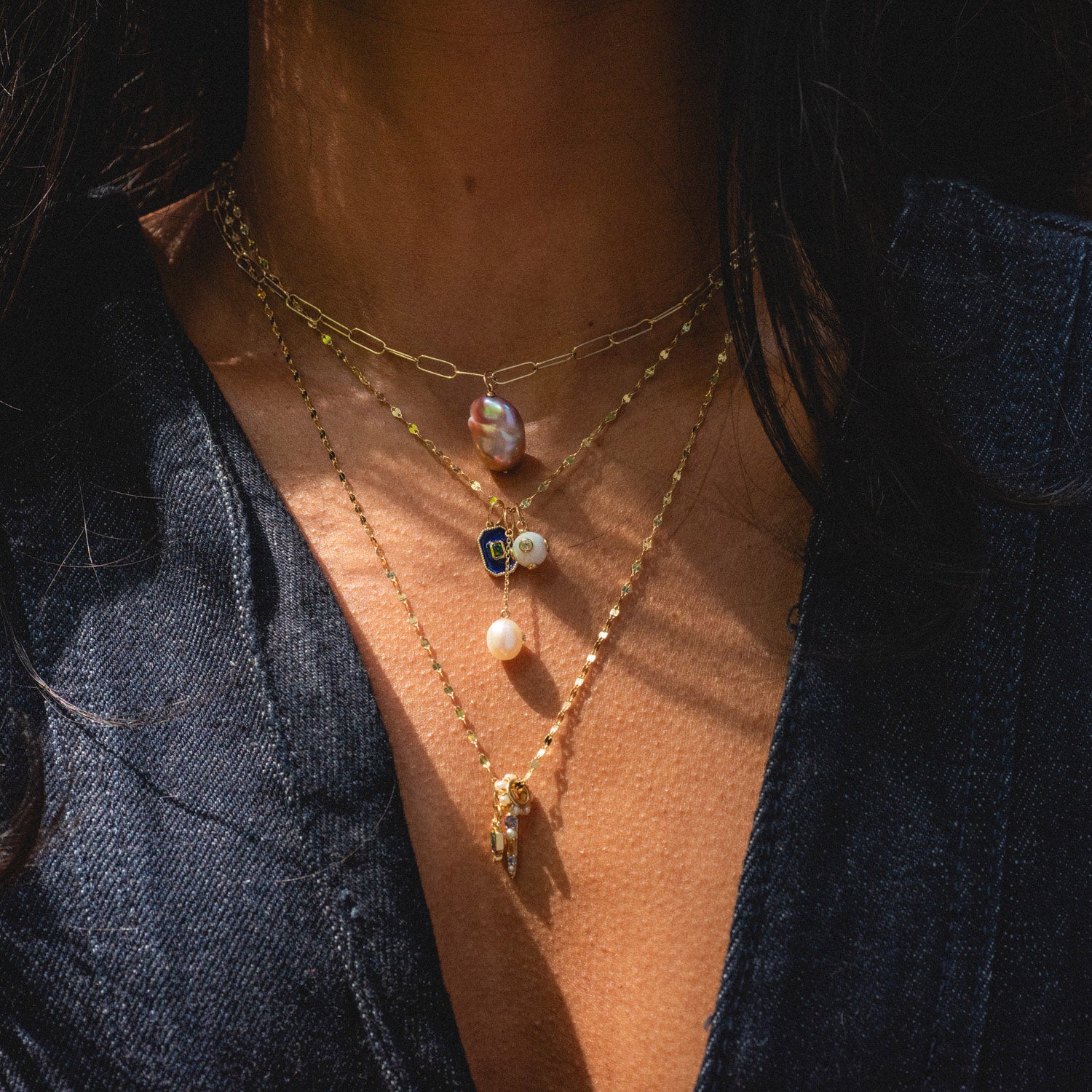 A person in a dark denim top wears three layered gold necklaces with unique charms—a pearl, a blue gem, and the Solid Gold Statement Charm—highlighted by sunlight. All are Local Eclectic exclusives.