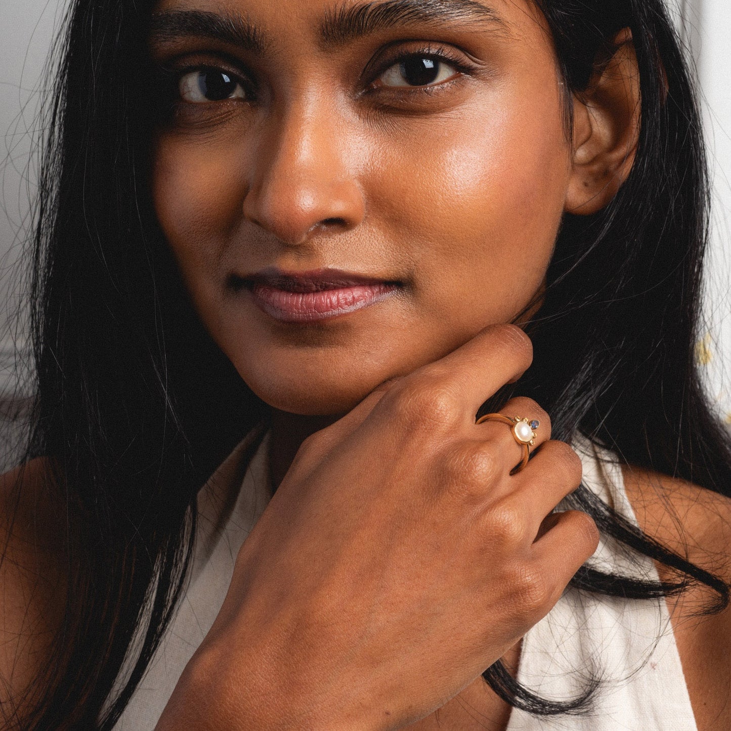 A woman with long dark hair gazes at the camera, resting her chin on her hand and wearing the Solid Gold Crowned Bezel Pearl Ring. She has smooth skin, a slight smile, and is dressed in a light-colored top.
