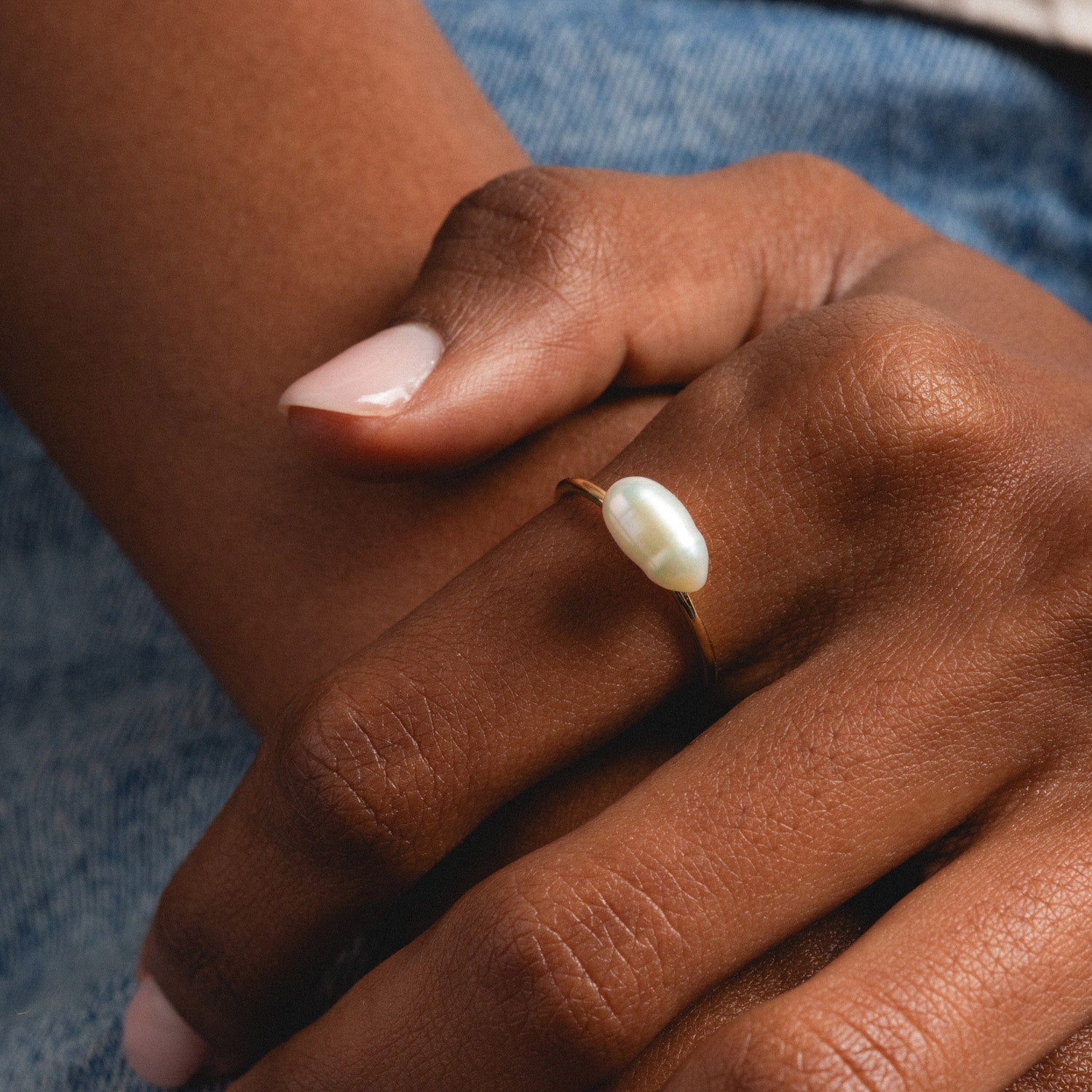 A close-up of a hand with manicured nails wearing the Solid Gold Organic Pearl Solitaire Ring, featuring a single white pearl, resting on denim fabric.