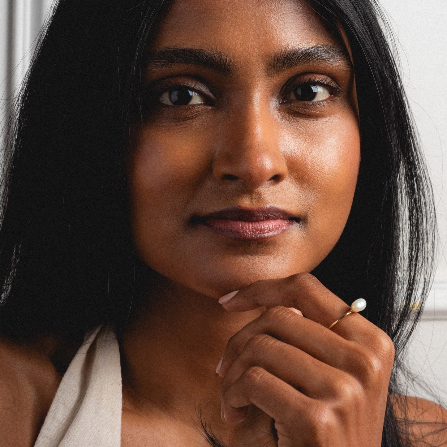 A woman with long dark hair and smooth brown skin looks at the camera, chin resting on her hand. She wears the Solid Gold Organic Pearl Solitaire Ring and a light-colored top, with a softly blurred background.