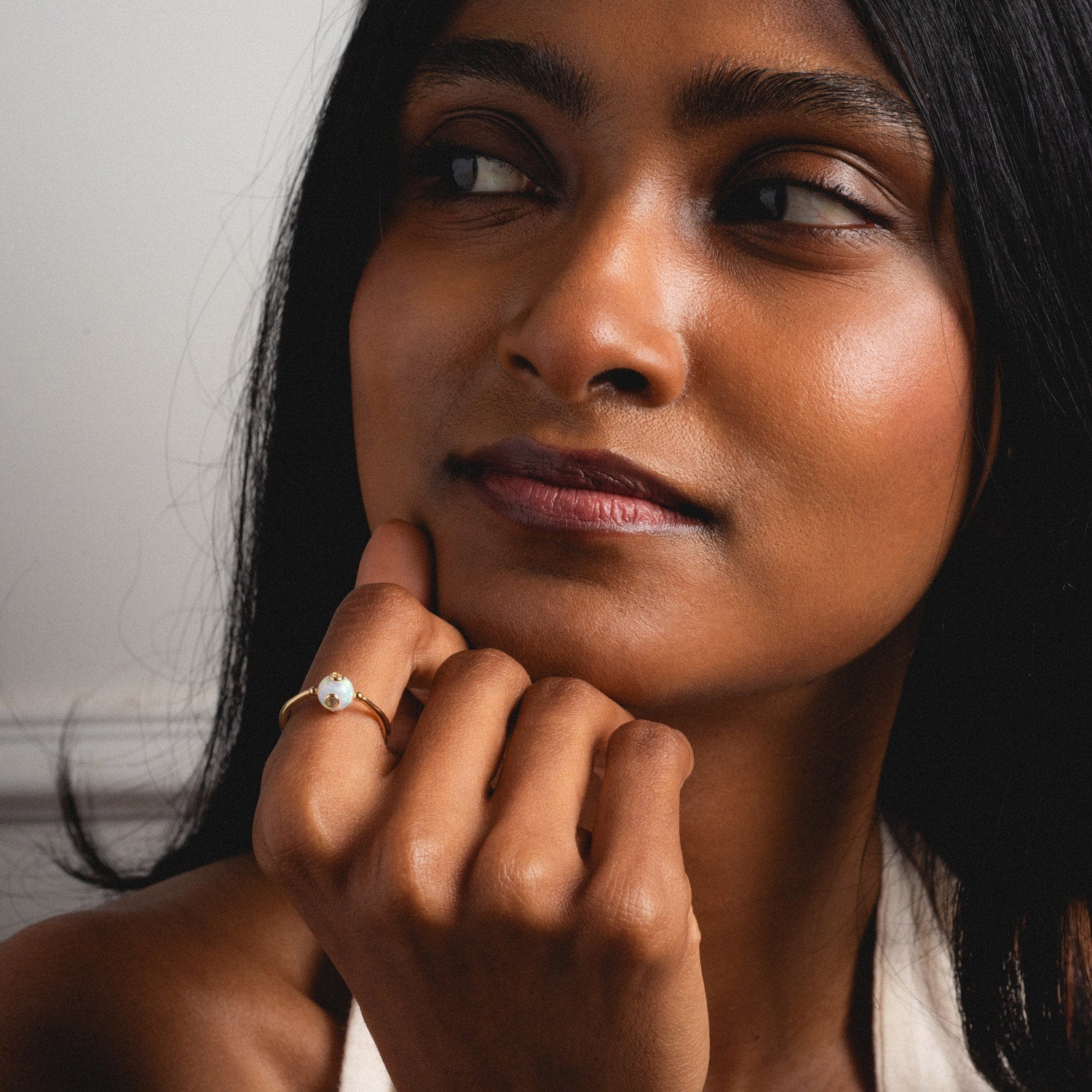 A woman with long dark hair gazes thoughtfully to the side, her chin on her hand, wearing the Solid Gold Gem Drop Opal Spinner Ring with a round white opal stone on her finger.