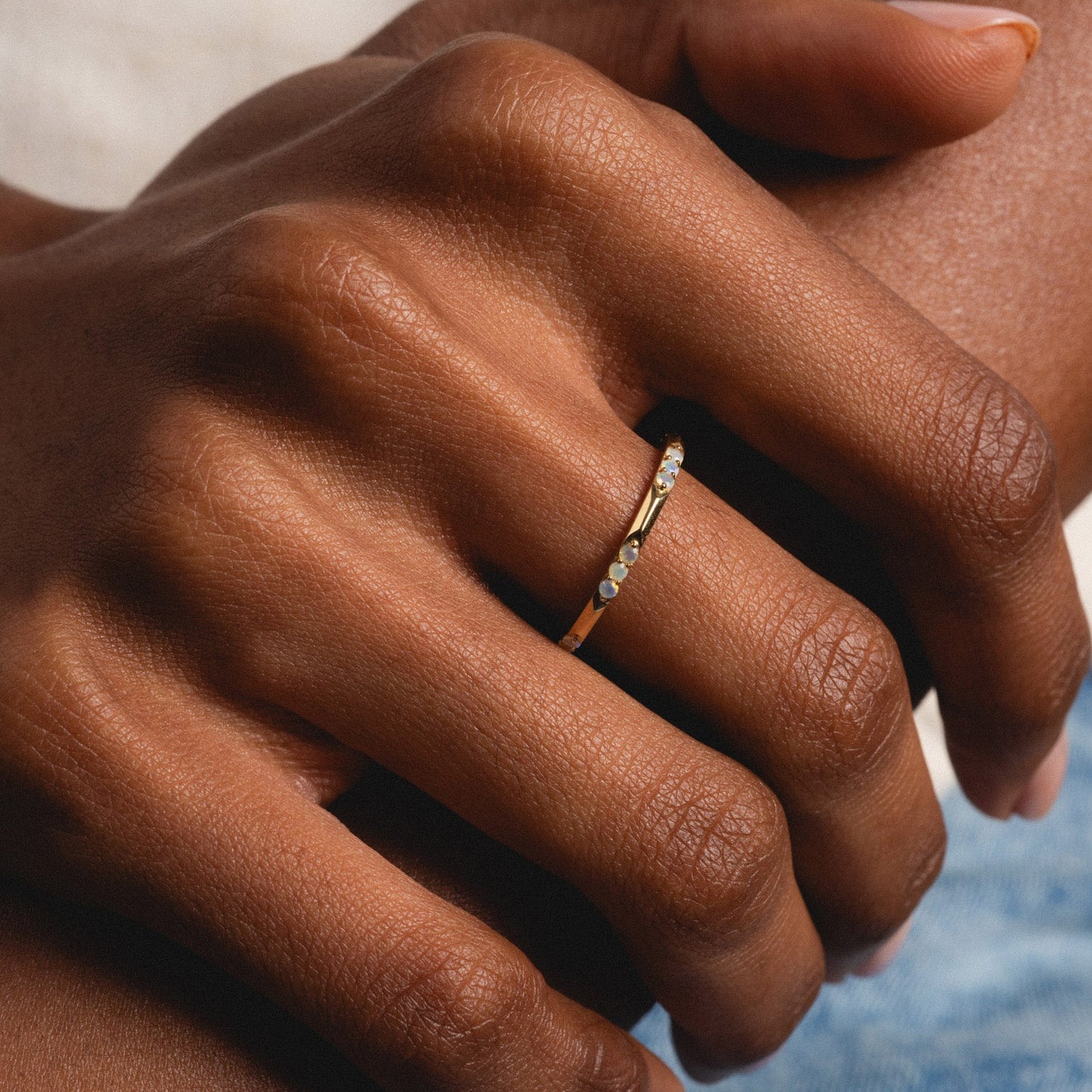 A close-up of a hand with medium-brown skin tone wearing the Solid Gold Beveled Opal Stacking Ring on the ring finger, gently resting on another hand.