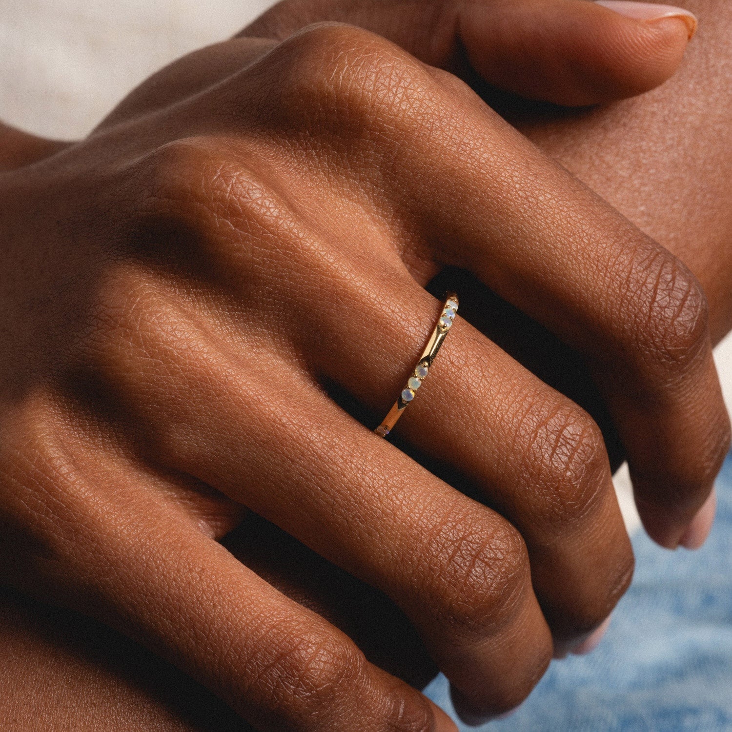 A close-up of a hand with medium-brown skin tone wearing the Solid Gold Beveled Opal Stacking Ring on the ring finger, gently resting on another hand.