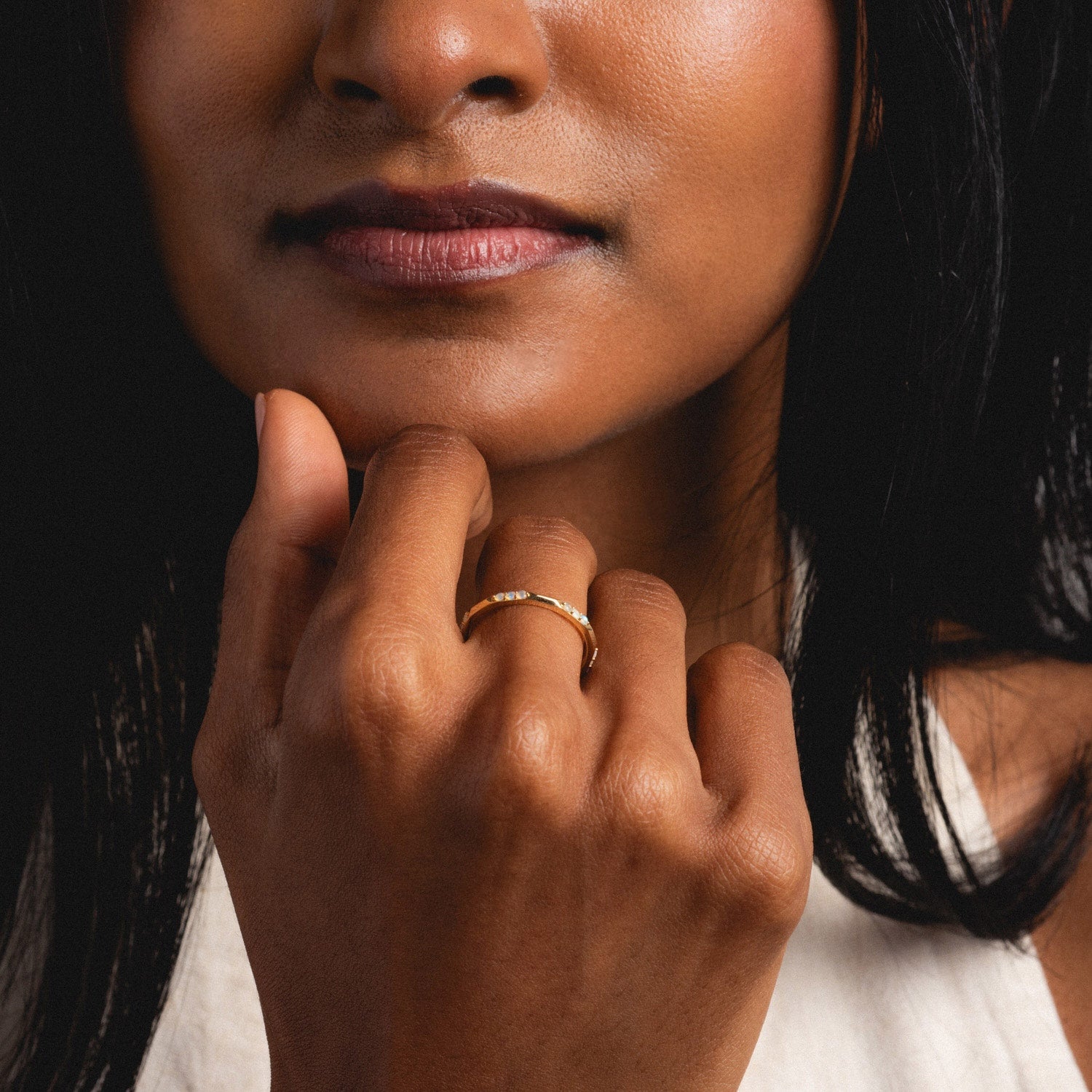 A woman with medium brown skin and dark hair wears the Solid Gold Beveled Opal Stacking Ring on her finger, gently touching her chin. She has natural makeup, a light-colored top, and the focus is on her hand and lips.