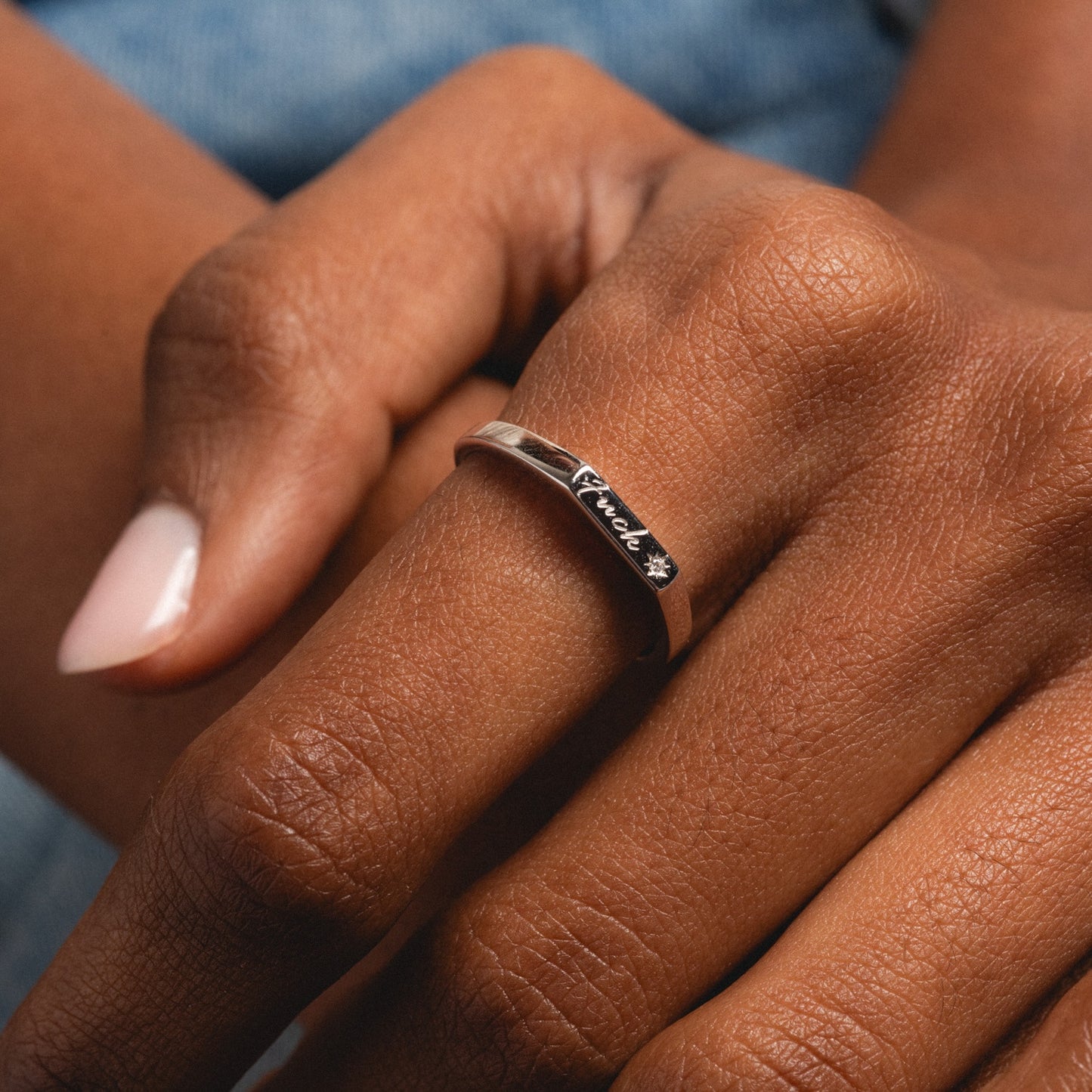 A close-up of a hand with medium brown skin wearing the Local Eclectic exclusive Tiny Fuck Ring in silver, engraved with “Friend” and a small star. The neatly manicured nails are painted light pink.