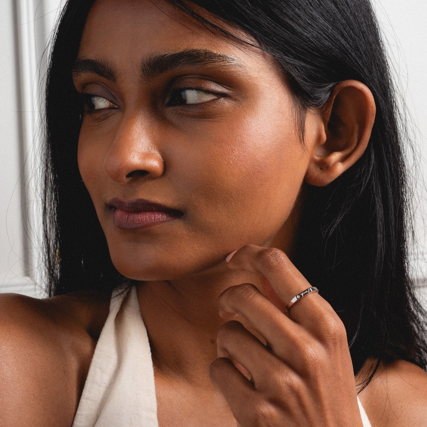 A woman with long dark hair and a neutral expression looks to the side, wearing a sleeveless top and the Tiny Fuck Ring in 14k yellow gold plate, gently touching her chin with her hand as she shows off the bold accessory.