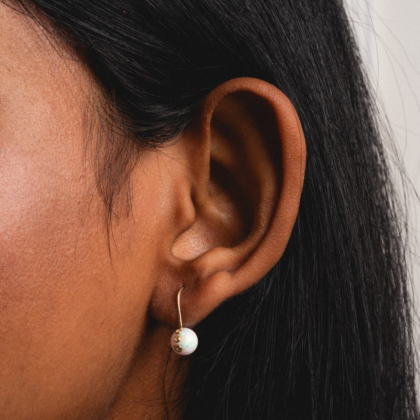 A close-up of a person with smooth, dark brown skin and straight black hair wearing Solid Gold Floating Opal Gem Drop Earrings featuring a round white opal.