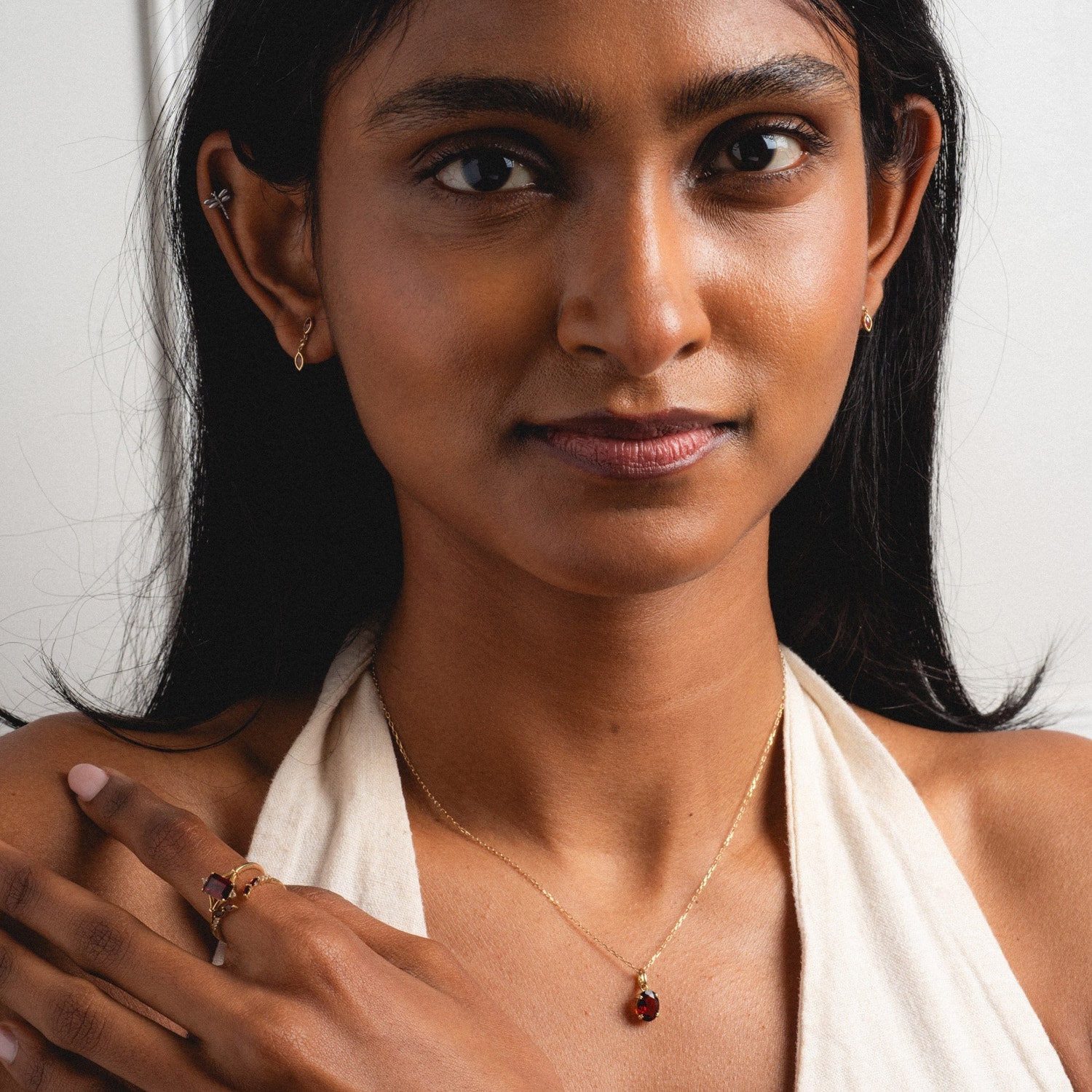 A woman with long dark hair wears the Solid Gold 2025 January Capsule Garnet Full Set, featuring a cream halter top, gold earrings, necklace with a red garnet pendant, and rings. She gazes at the camera, one hand resting on her shoulder.