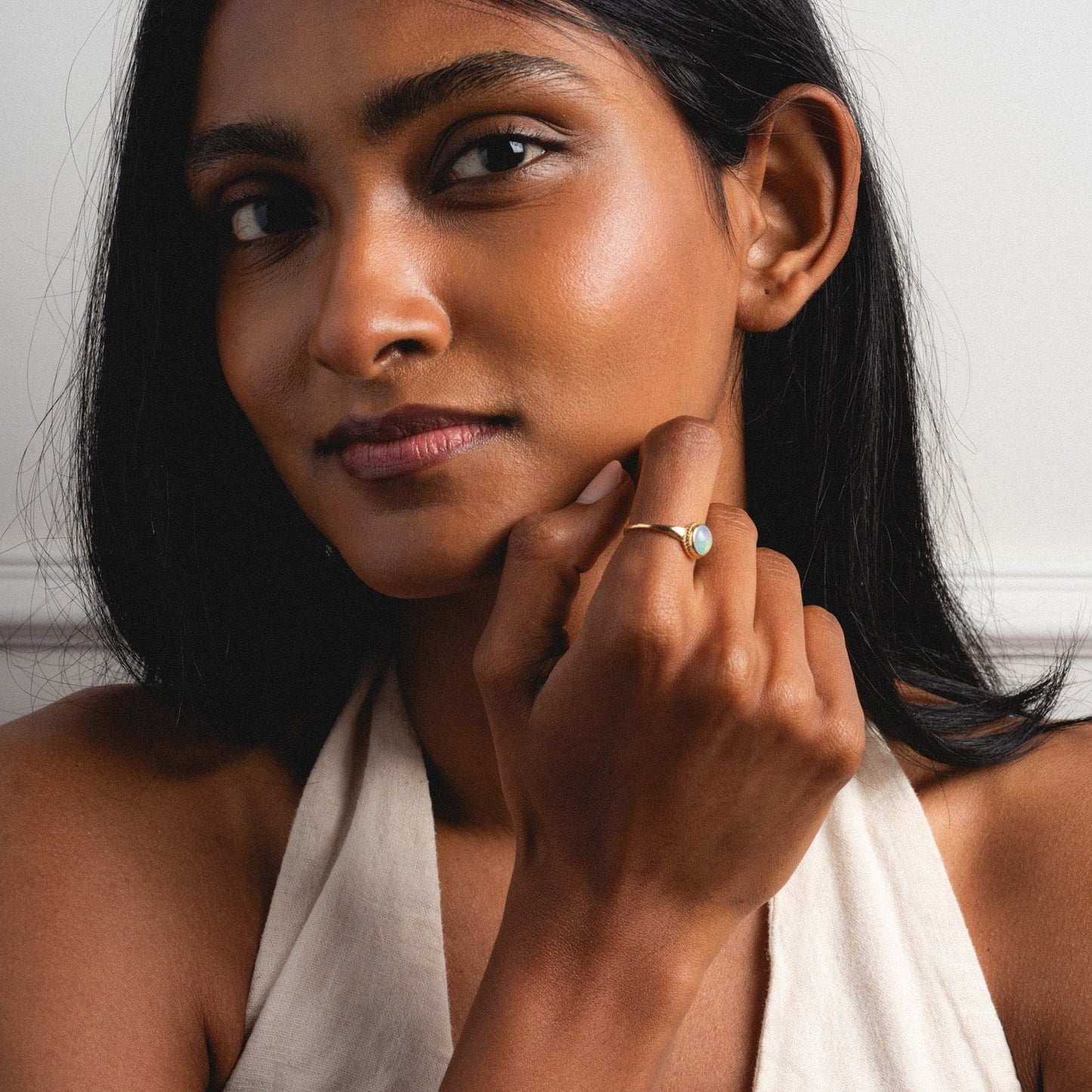 A woman with long dark hair and a calm, confident expression wears a sleeveless beige top as she showcases the Solid Gold Textured Opal Signet Ring near her chin, highlighting its light-colored stone for the camera.