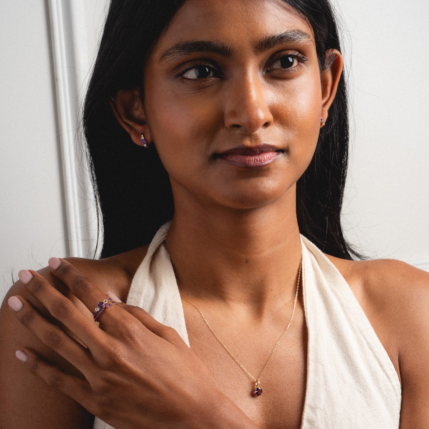 A woman with long dark hair wears the Solid Gold 2025 February Capsule Garnet Full Set, featuring a cream halter top, gold necklace and matching earrings with purple garnet stones, plus a ring. She gazes calmly to the side. Includes free gift.