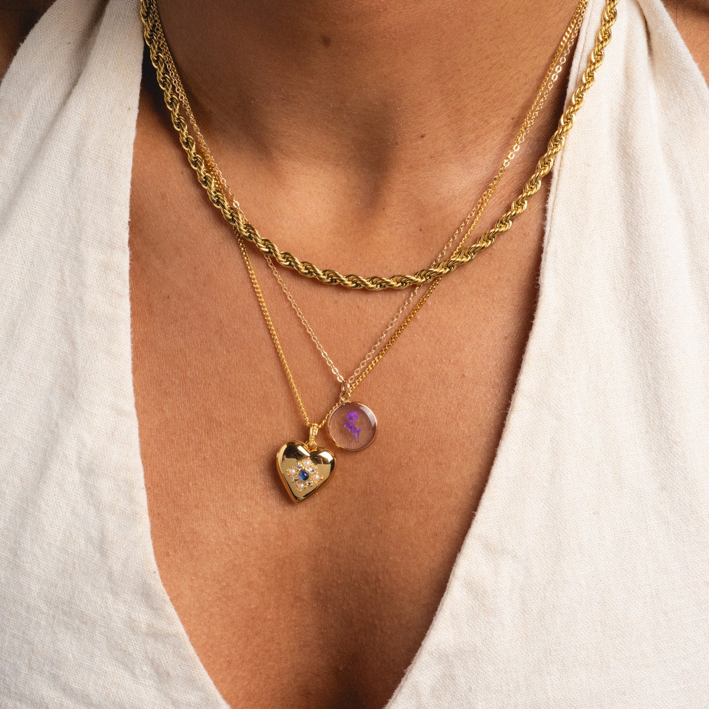 A close-up of a person wearing three gold necklaces over a white top, including the Tiny Birth Flower Portrait Necklace with two pendants: a gold heart set with a blue gem and a round pendant featuring a purple birth flower design.