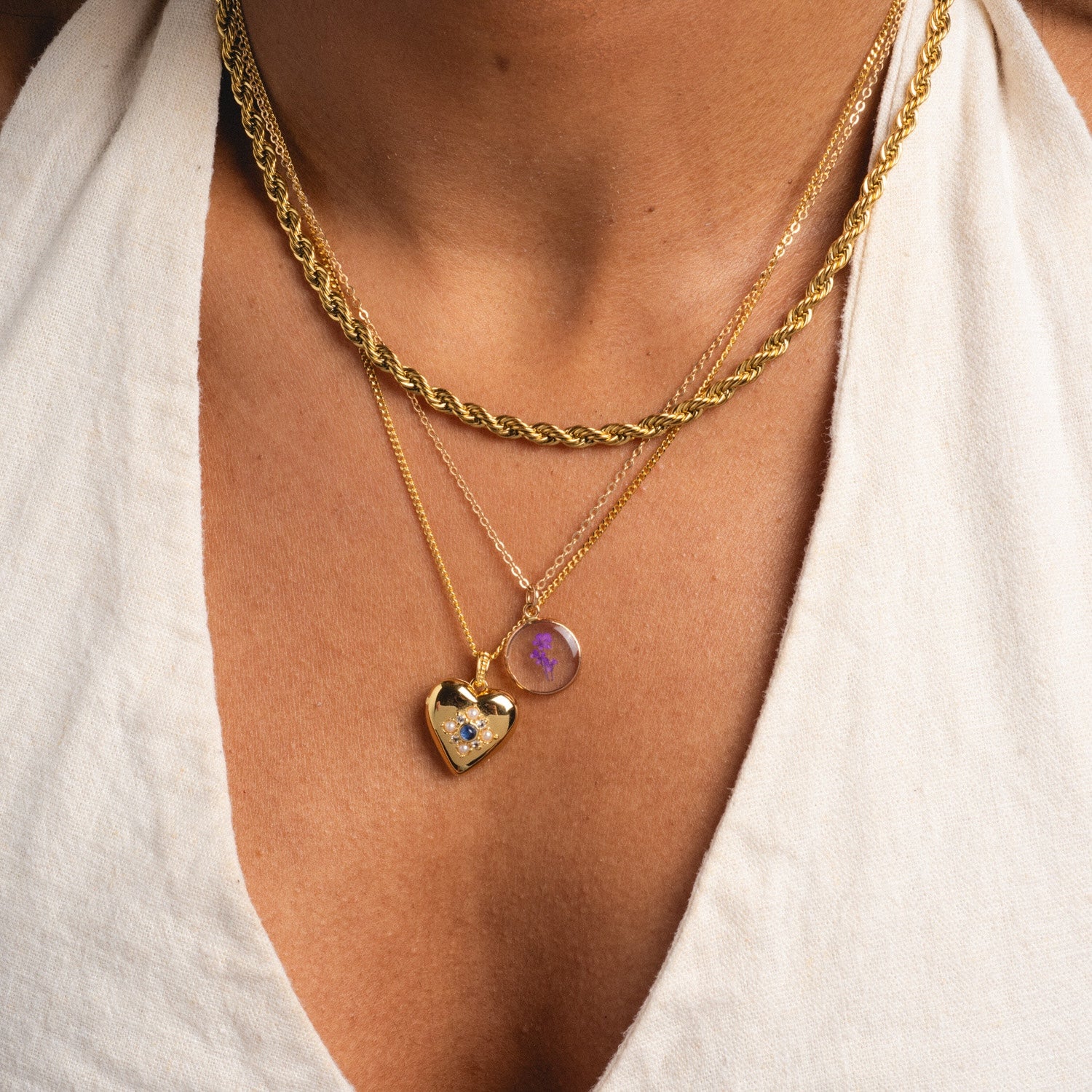 A close-up of a person wearing three gold necklaces over a white top, including the Tiny Birth Flower Portrait Necklace with two pendants: a gold heart set with a blue gem and a round pendant featuring a purple birth flower design.