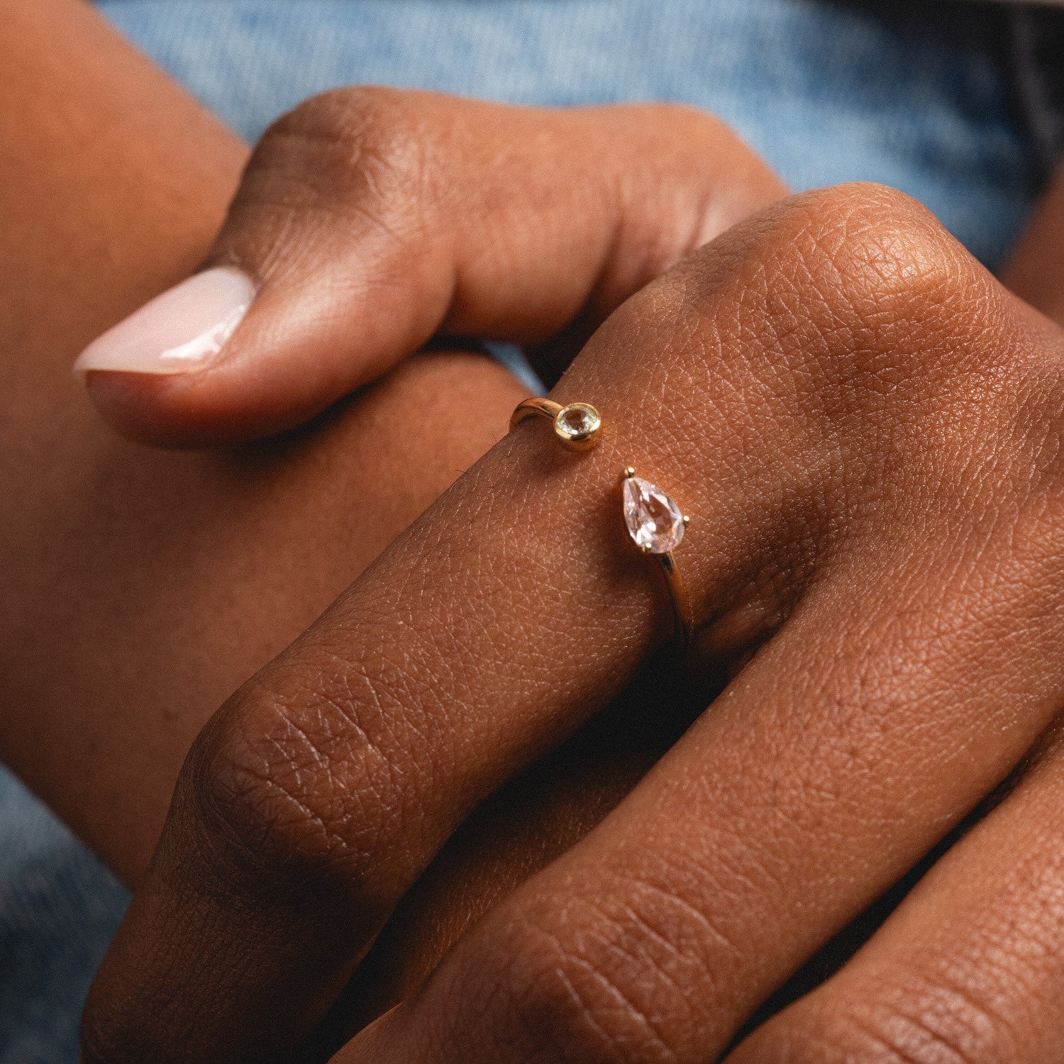 A close-up of a hand wearing the Solid Gold Sapphire and Aquamarine Open Front Ring, featuring a small round sapphire on one end and a teardrop-shaped aquamarine on the other, against a blurred denim background.