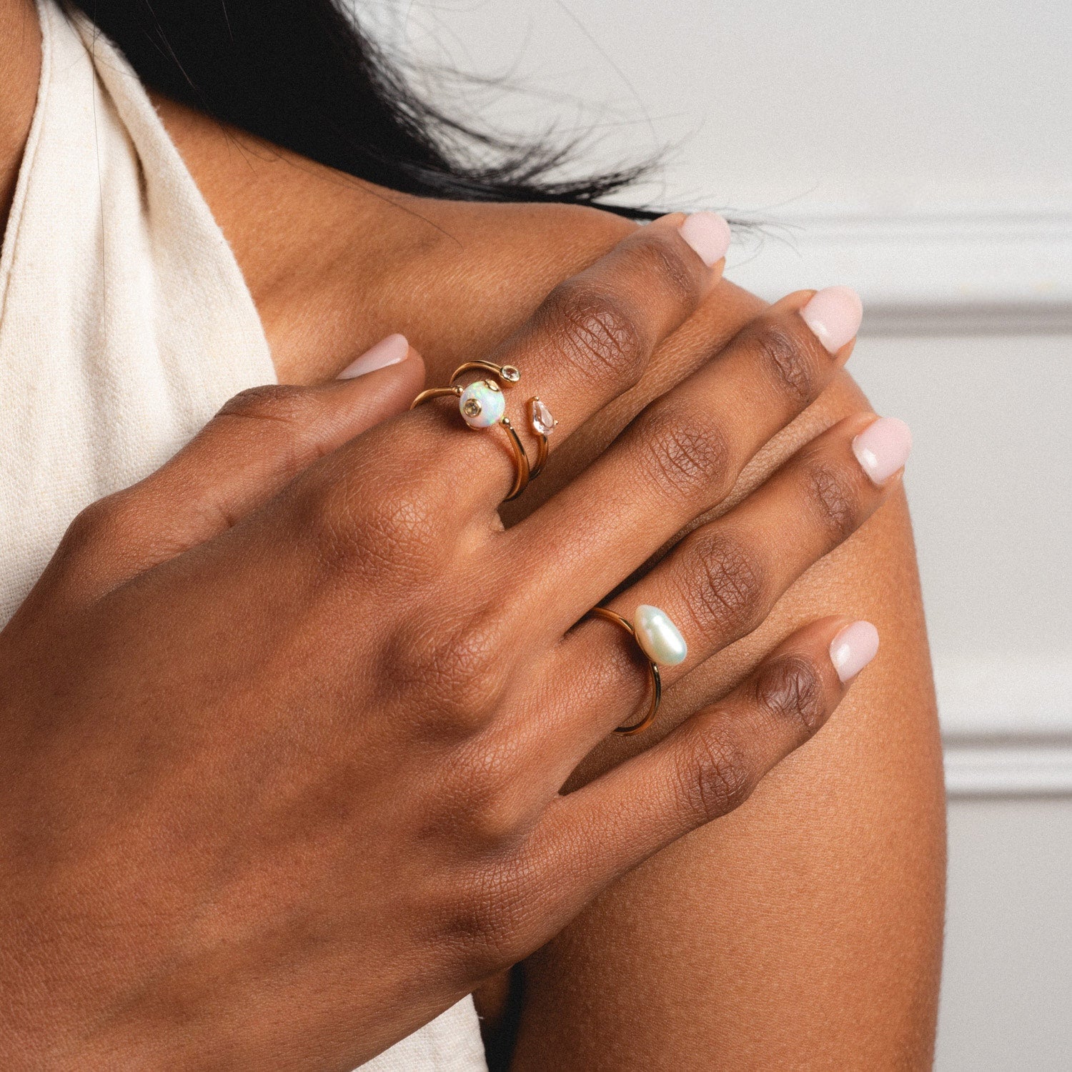 A close-up of a woman's manicured hands wearing several gold rings, including the Solid Gold Gem Drop Opal Spinner Ring, some with pearls and gemstones, resting gently on her shoulder.