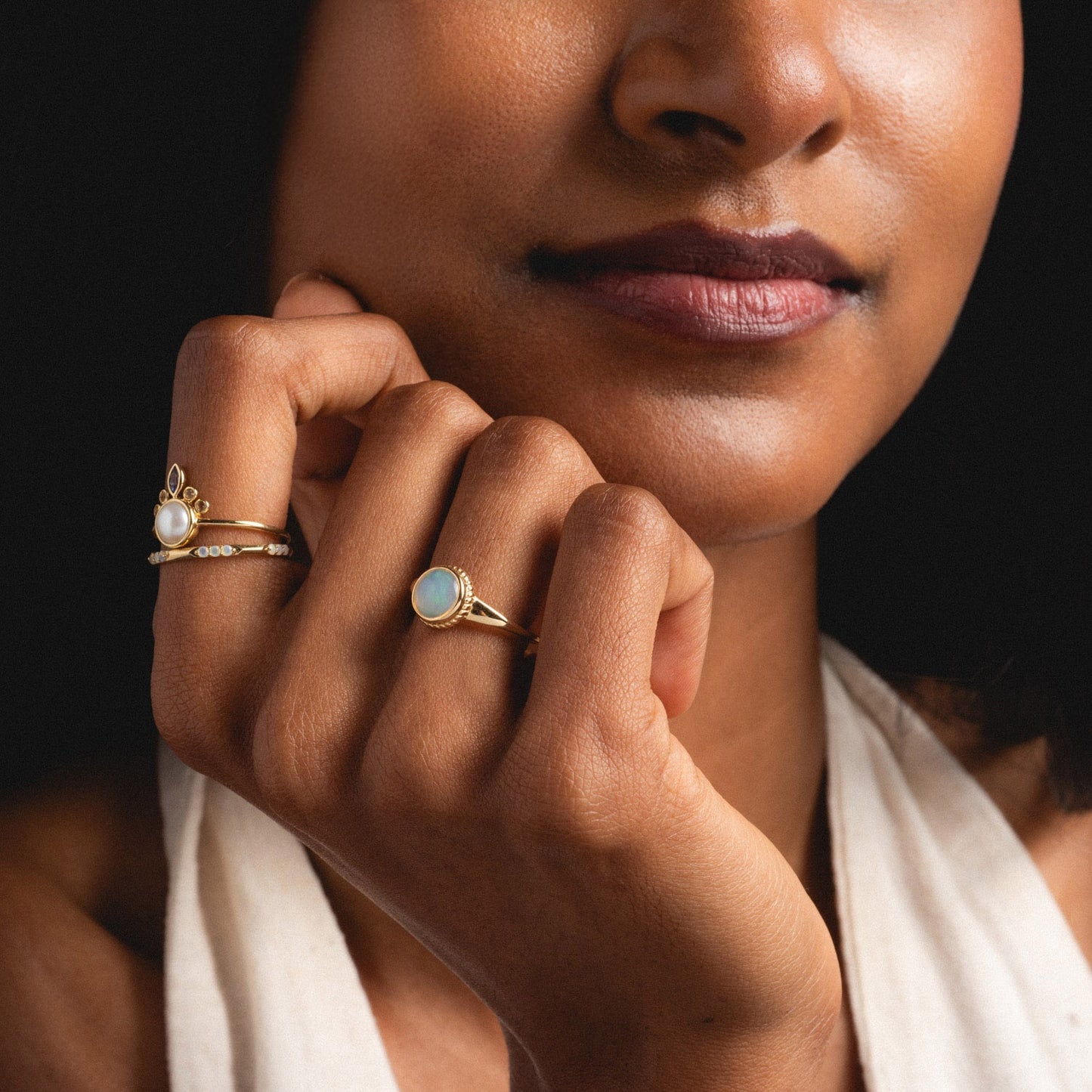 A woman with medium brown skin touches her face, showcasing the Solid Gold Textured Opal Signet Ring on her finger. She has a natural makeup look and wears a light halter top, with the focus on her hand and jewelry.