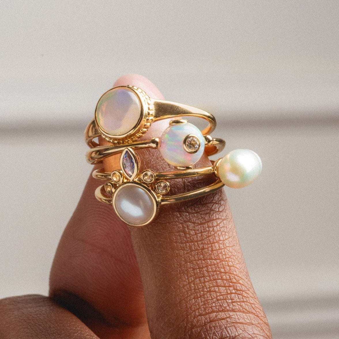 A hand with dark skin tone wears four Solid Gold Gem Drop Opal Spinner Rings, each adorned with pearls and opal-like stones, against a neutral background.