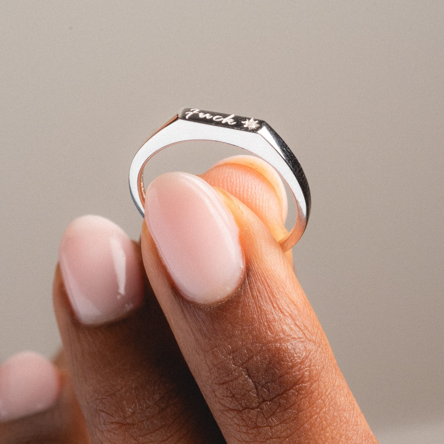 A hand with neatly manicured nails displays the Tiny Fuck Ring—its flat top engraved with “fuck” and a small star, finished in 14k yellow gold plating, set against a neutral background.