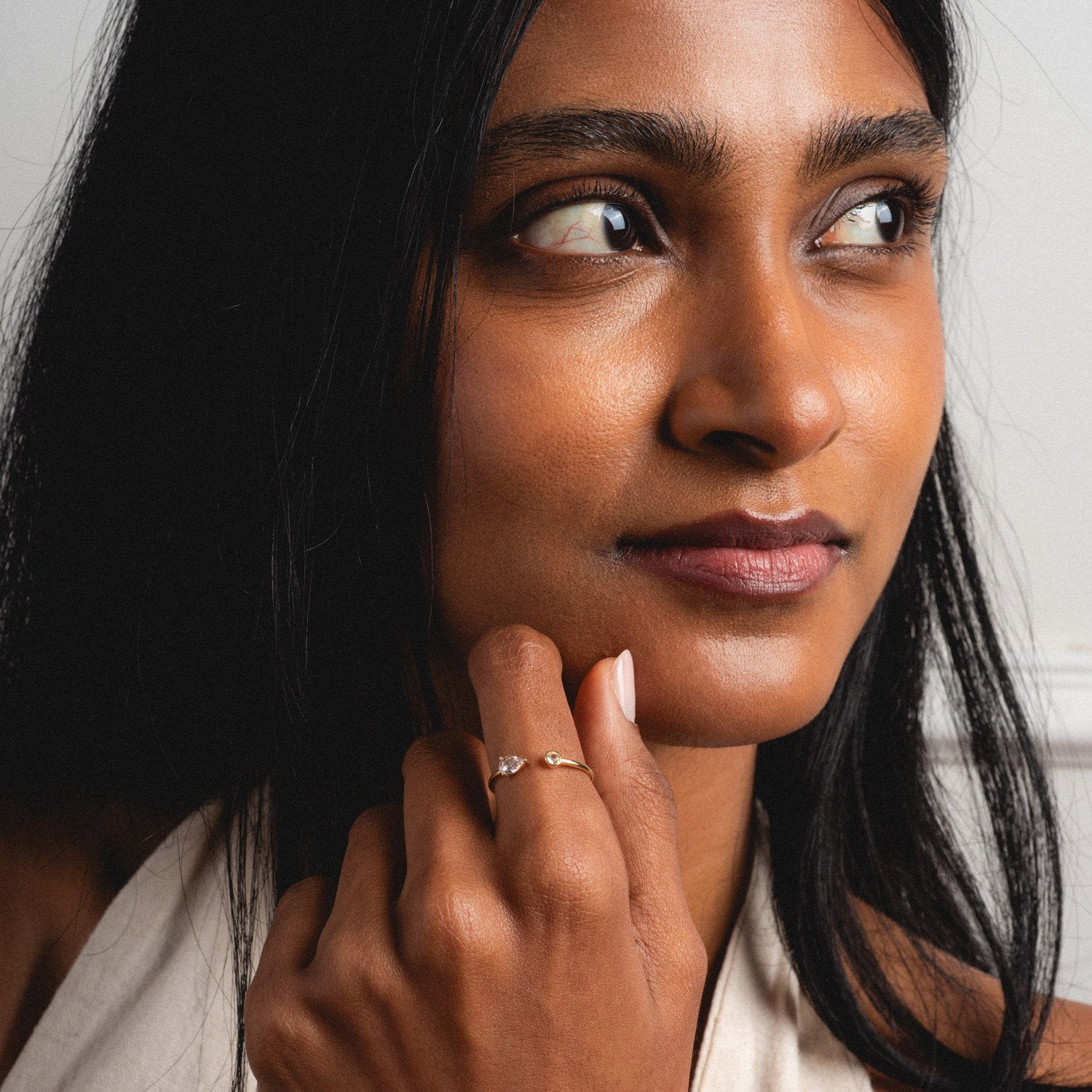 A woman with long dark hair gazes thoughtfully to the side, her hand near her face displaying the Solid Gold Sapphire and Aquamarine Open Front Ring. She has smooth skin and wears a light-colored top.