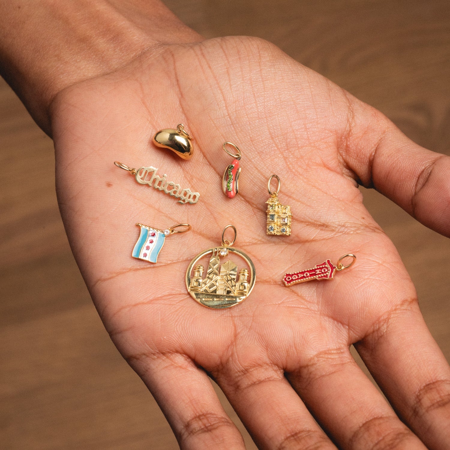 A hand displays the Solid Gold Chicago Charm alongside six others—a red apple, “Chicago” text, a house, city flag, circular cityscape, skyline, and red arrow—all set against a wooden background.