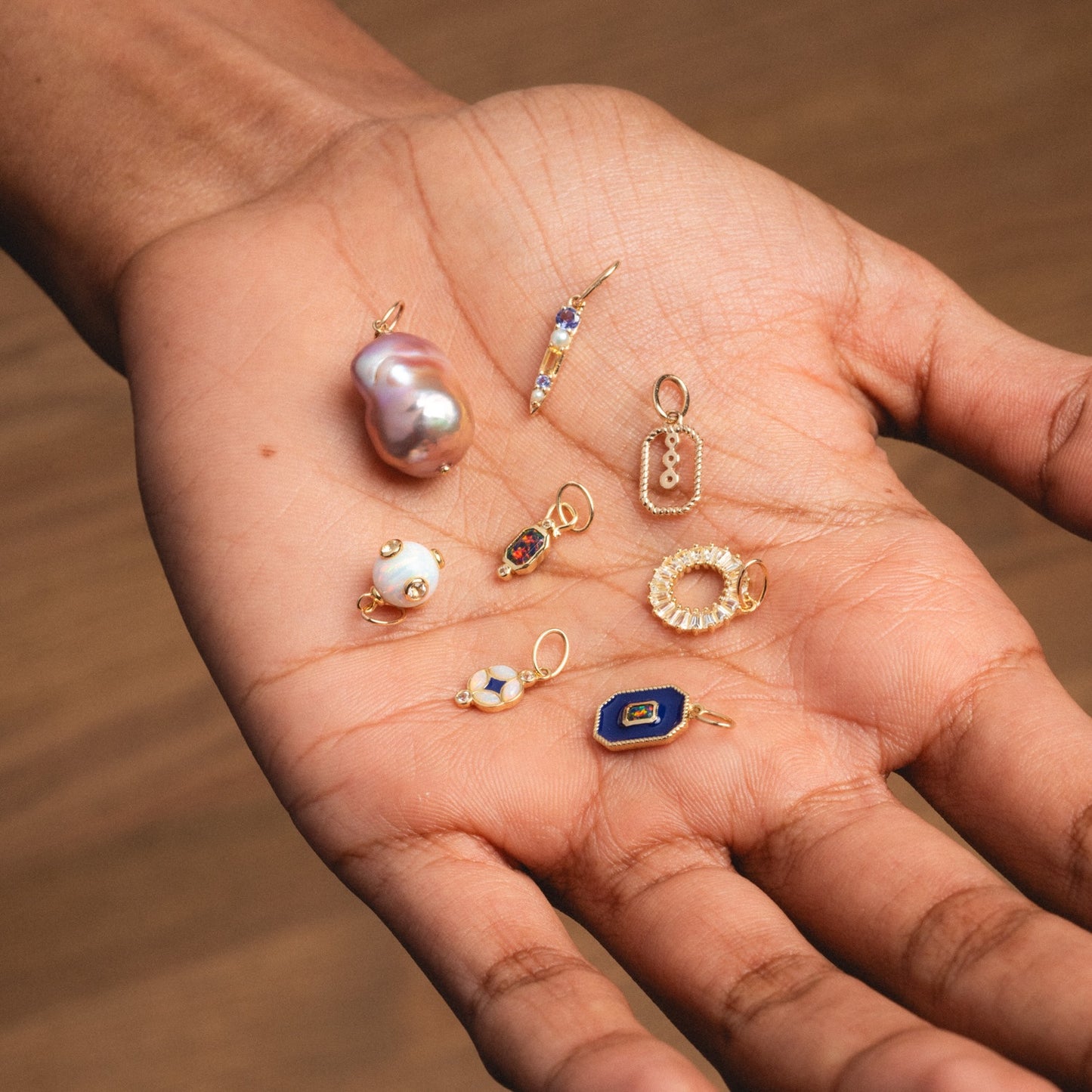 A hand displays several small, decorative charms—pearls, beads, gemstones, and the Solid Gold Statement Charm—arranged neatly on the palm against a brown background. Some pieces are exclusive designs from Local Eclectic.