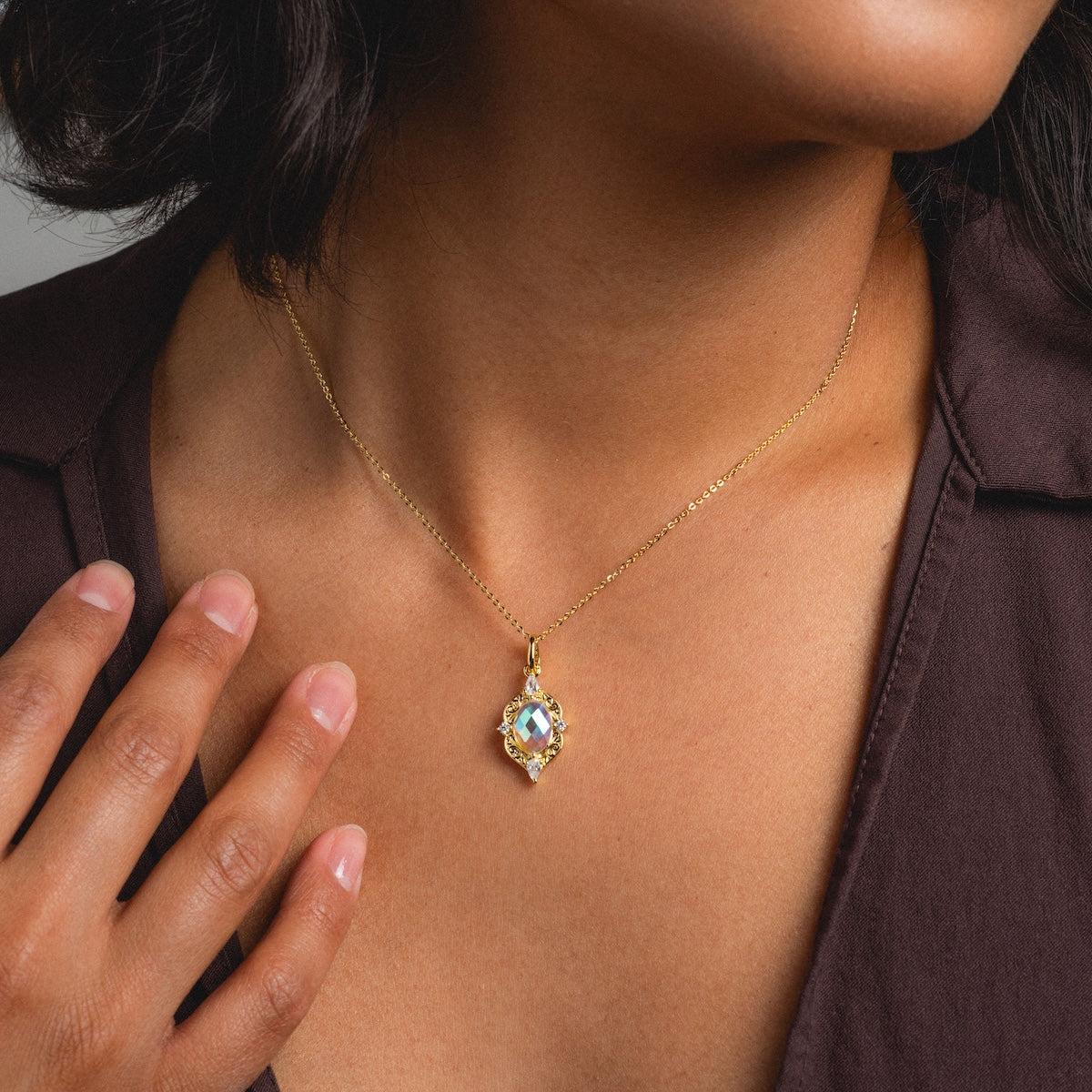 A person wearing the Lady Luxe Angel Aura Quartz Pendant Necklace, featuring a delicate 14k gold-plated chain with a light blue gemstone pendant, poses in a dark open-collar shirt with one hand near the necklace.