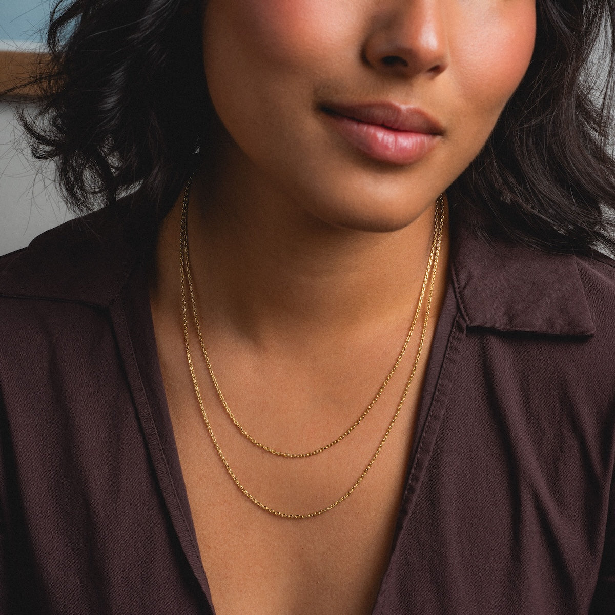 A woman in a dark brown collared shirt wears two delicate gold necklaces, including the Oval Link Chain. Her wavy dark hair and natural makeup complement her subtle smile.