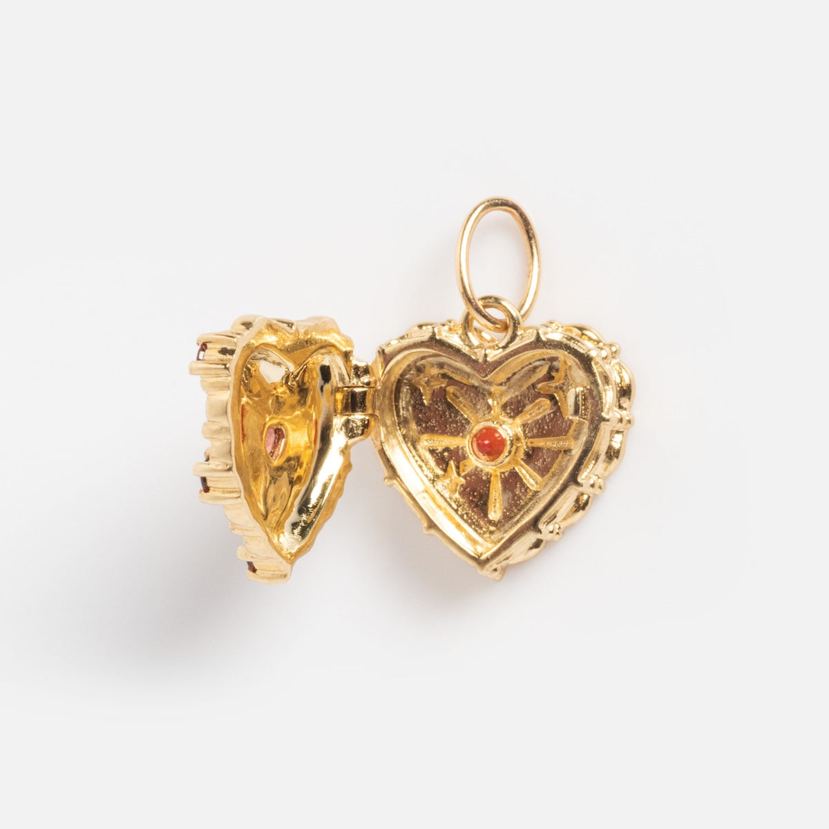 The Solid Gold Celebration Charm is a 9k gold heart-shaped locket with a textured edge, intricate interior design, and a small red gem at the center on each side. It includes a circular loop for easy attachment to your chain.