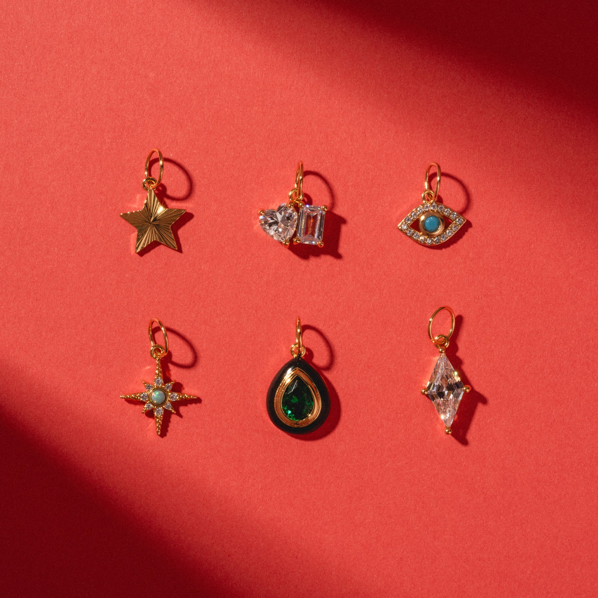 Six Charms by Jurate JLA, yellow gold plated with gemstone and simulated diamond accents, are arranged on a red background in star, heart, eye, starburst, teardrop, and diamond shapes.