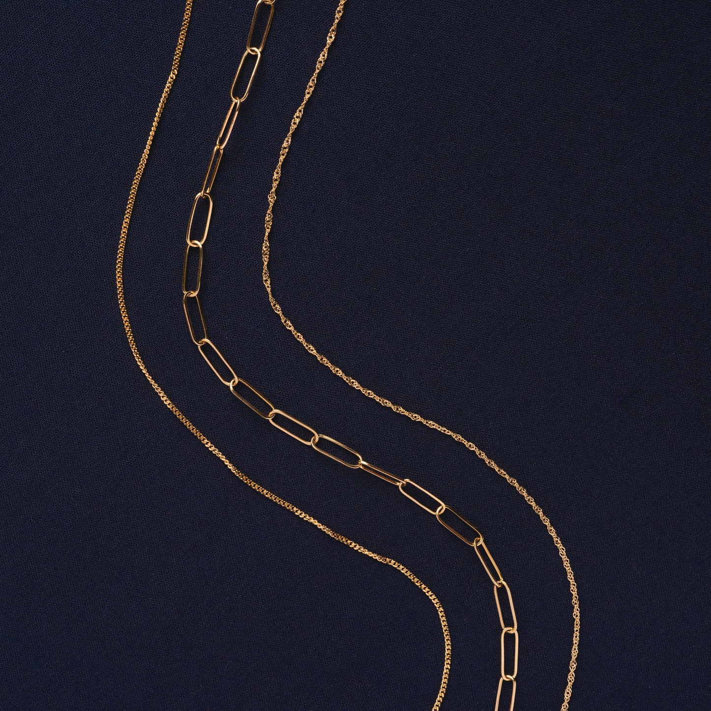 Three Solid Gold Perfect Start Charm Holder Necklaces, each with a unique chain design—paperclip, rope, and slim—are arranged in wavy lines on a dark blue textured background.