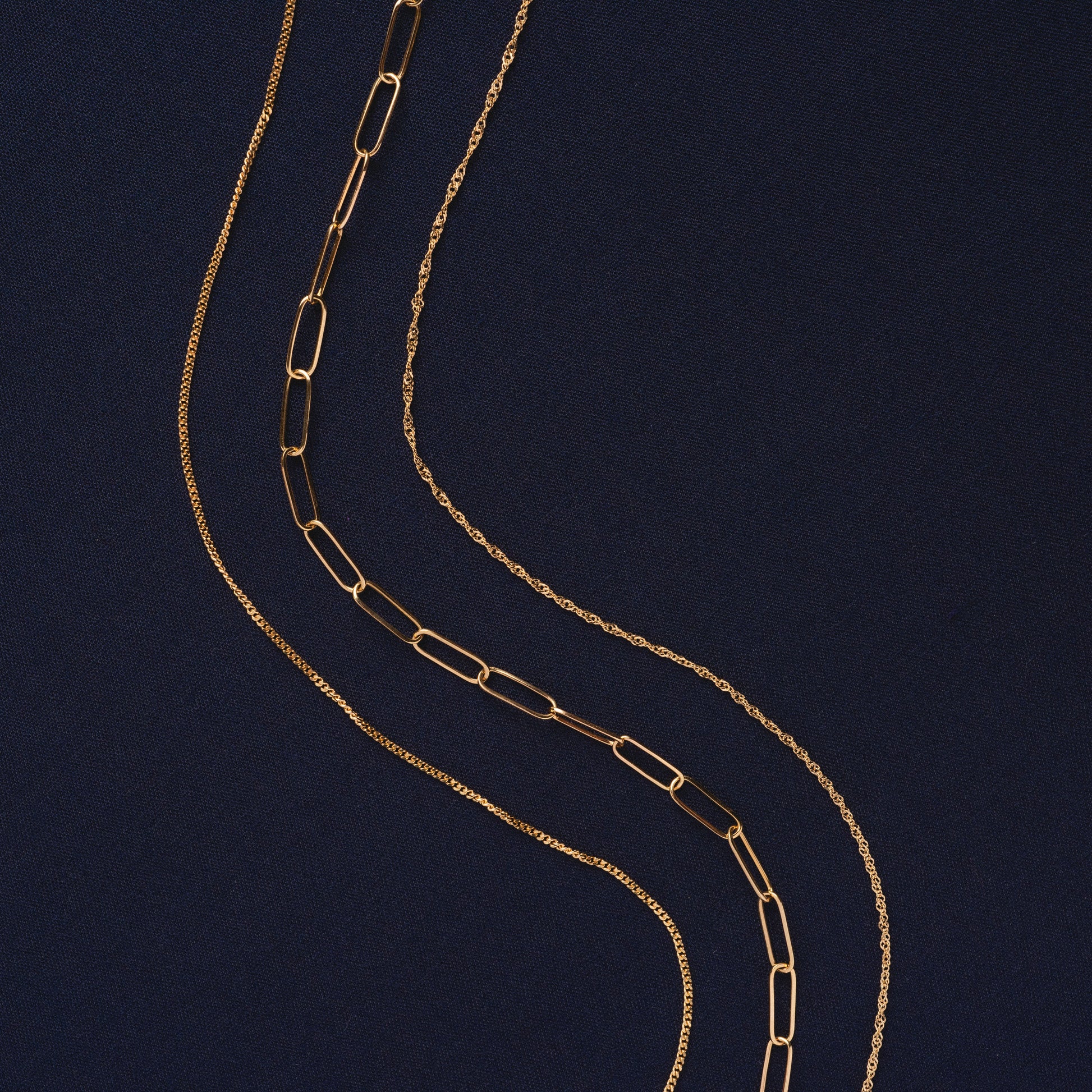 Three Solid Gold Perfect Start Charm Holder Necklaces, each with a unique chain design—paperclip, rope, and slim—are arranged in wavy lines on a dark blue textured background.