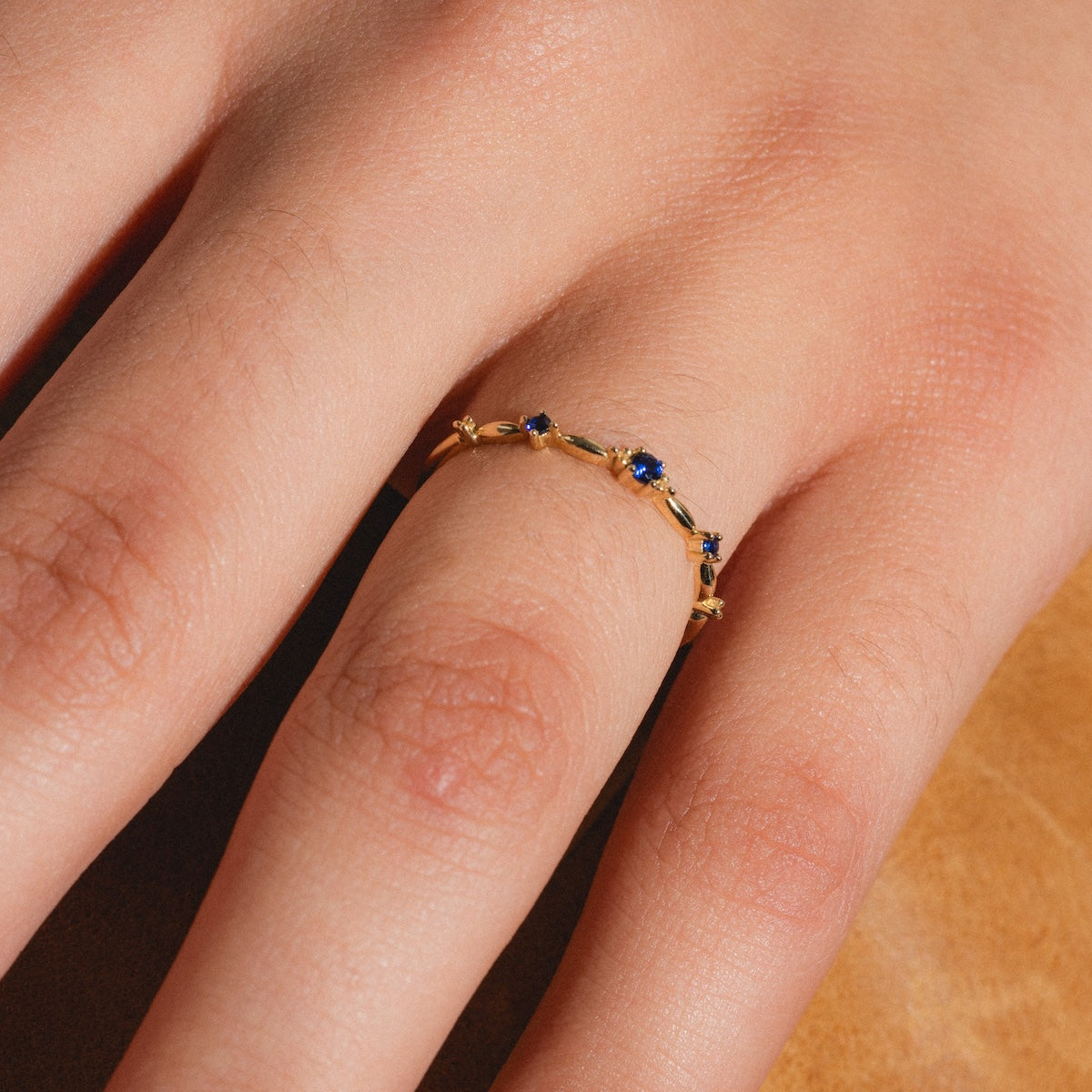 A hand wears the Solid Gold 2025 September Capsule Delicate Blue Sapphire Stacking Ring in 9k yellow gold with lab-created sapphires, shown close-up against a light brown background.
