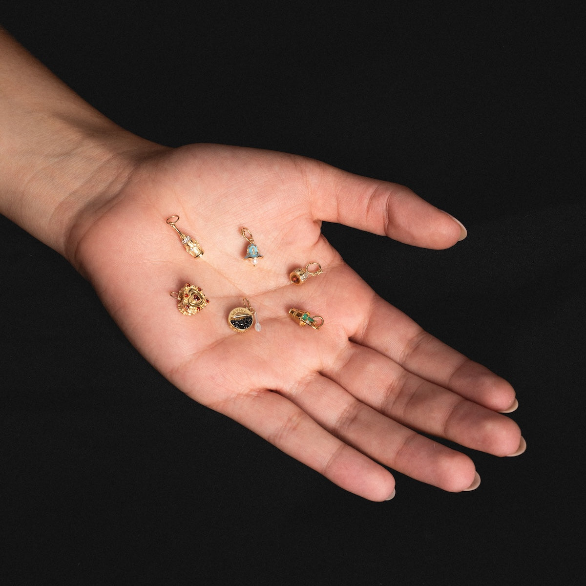 A hand with palm facing up displays six Solid Gold Celebration Charms crafted from 9k gold, each featuring colorful gemstones and intricate designs, all shown against a black background.