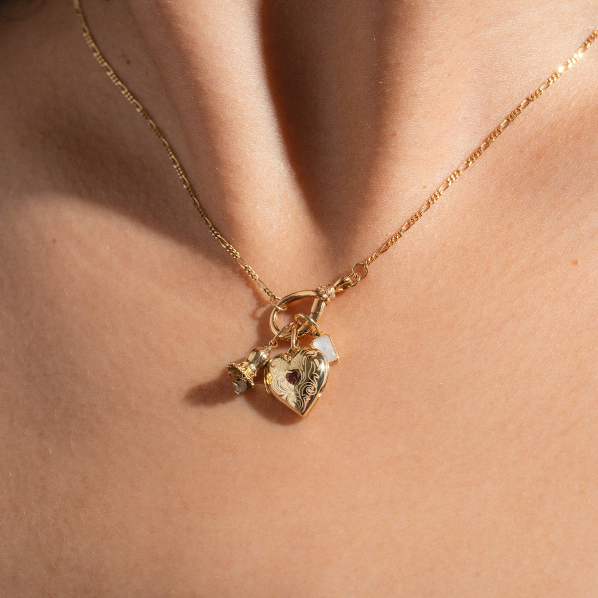 A close-up of the Solid Gold Figaro Charm Holder Necklace on a person’s neck, featuring a heart locket, rose charm, and clear gemstone charm on a delicate gold chain.