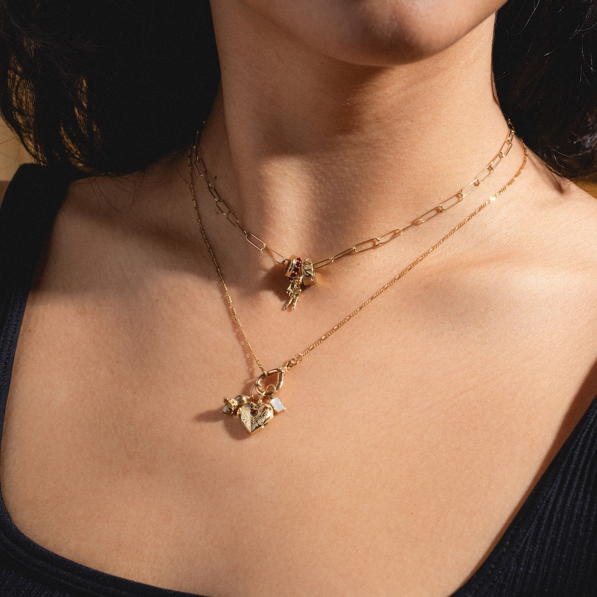 A person wearing the Solid Gold Figaro Charm Holder Necklace, featuring two gold chains—one with a small charm and one with a heart-shaped pendant—against bare skin and a black top.