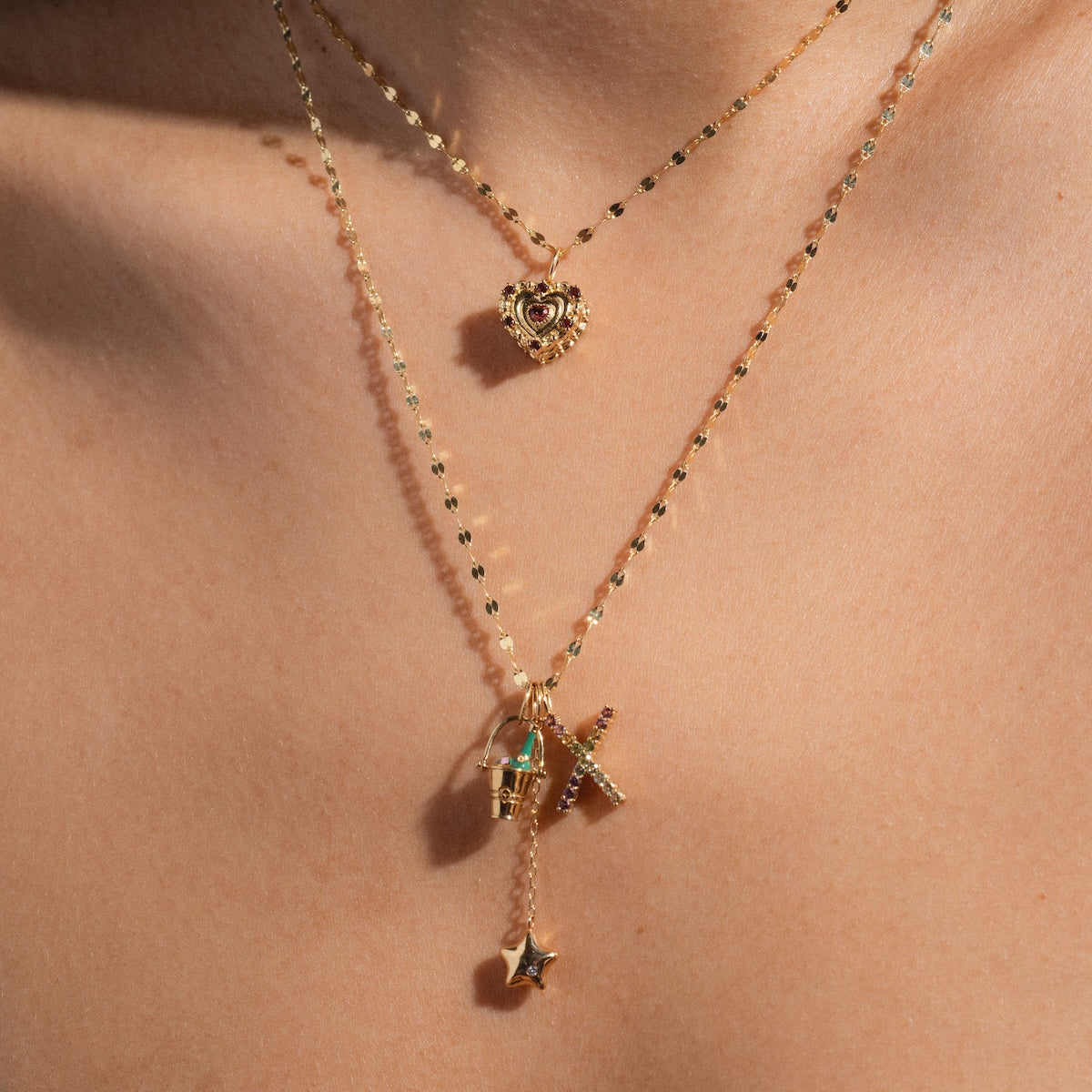 A close-up of a neck wearing two layered gold necklaces: the shorter with an ornate heart pendant, and the longer featuring the Solid Gold Celebration Charm—a 9k gold bucket, cross, and star—adding elegant detail.